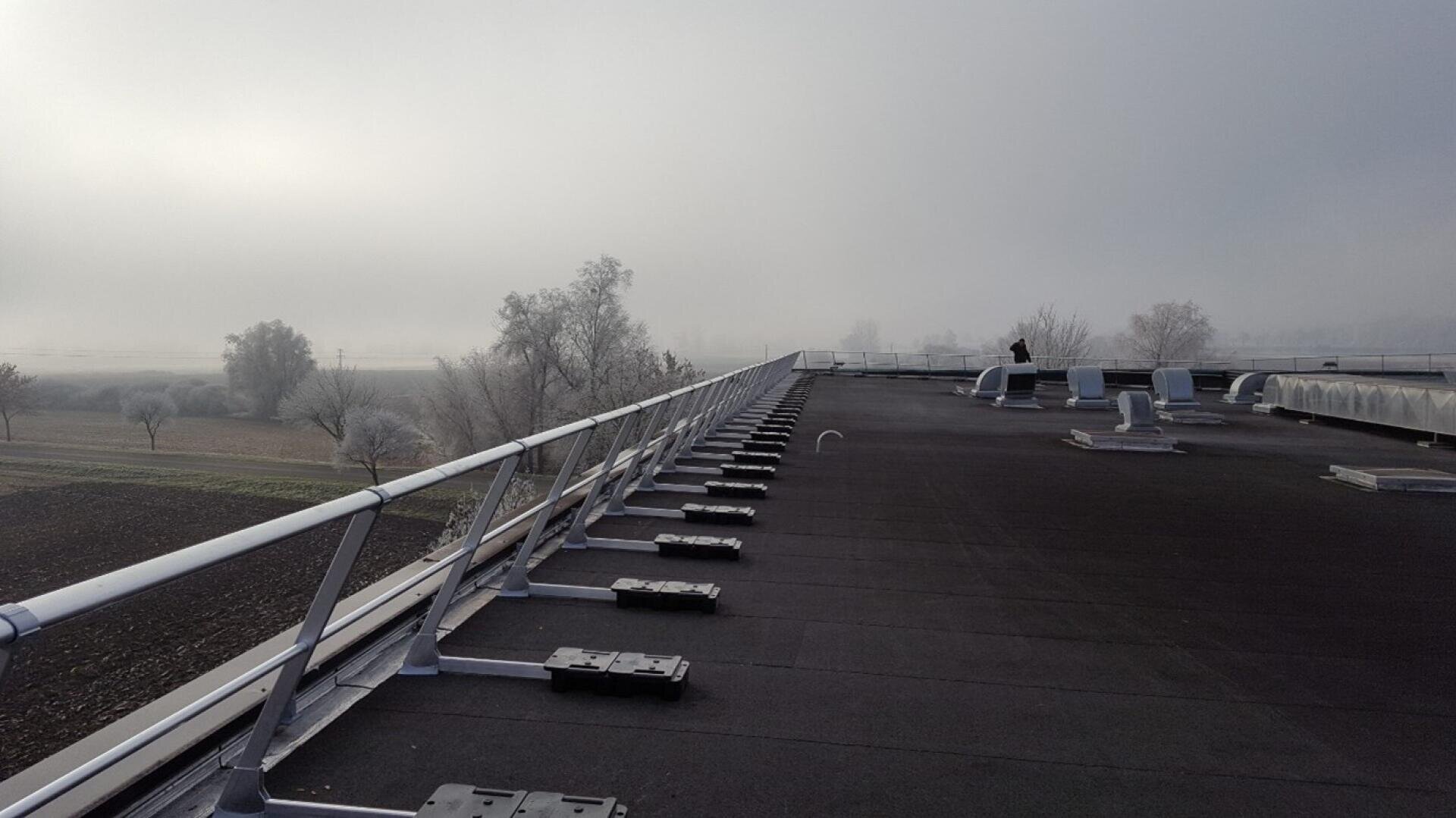 Ein flaches Dach mit Metallgeländern und Lüftungsanlagen, mit Blick auf eine neblige Landschaft mit kahlen Bäumen und Feldern. Eine Person steht in der Nähe des Randes in der Ferne, unter einem bedeckten Himmel.