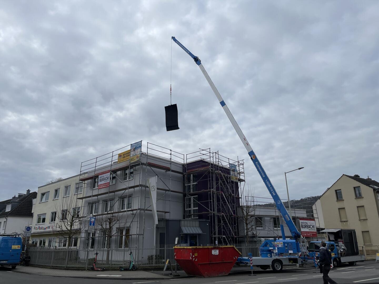 Ein Kran hebt ein großes schwarzes Objekt über ein eingerüstetes Gebäude, das sich an einem bewölkten Tag im Bau befindet. Auf der Straße sind mehrere Fahrzeuge und Baumaschinen zu sehen.