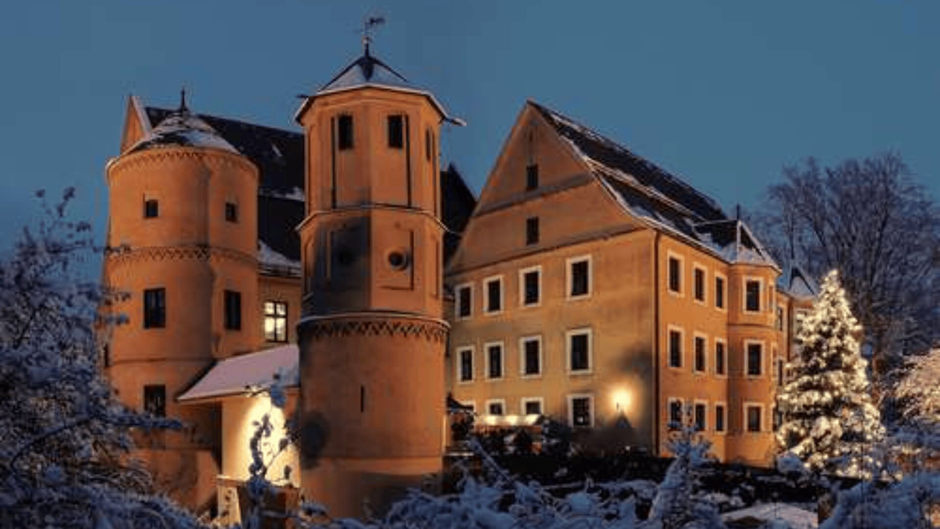 A warmly lit castle with round towers and steep roofs stands surrounded by snow-covered trees and a decorated Christmas tree in the evening.