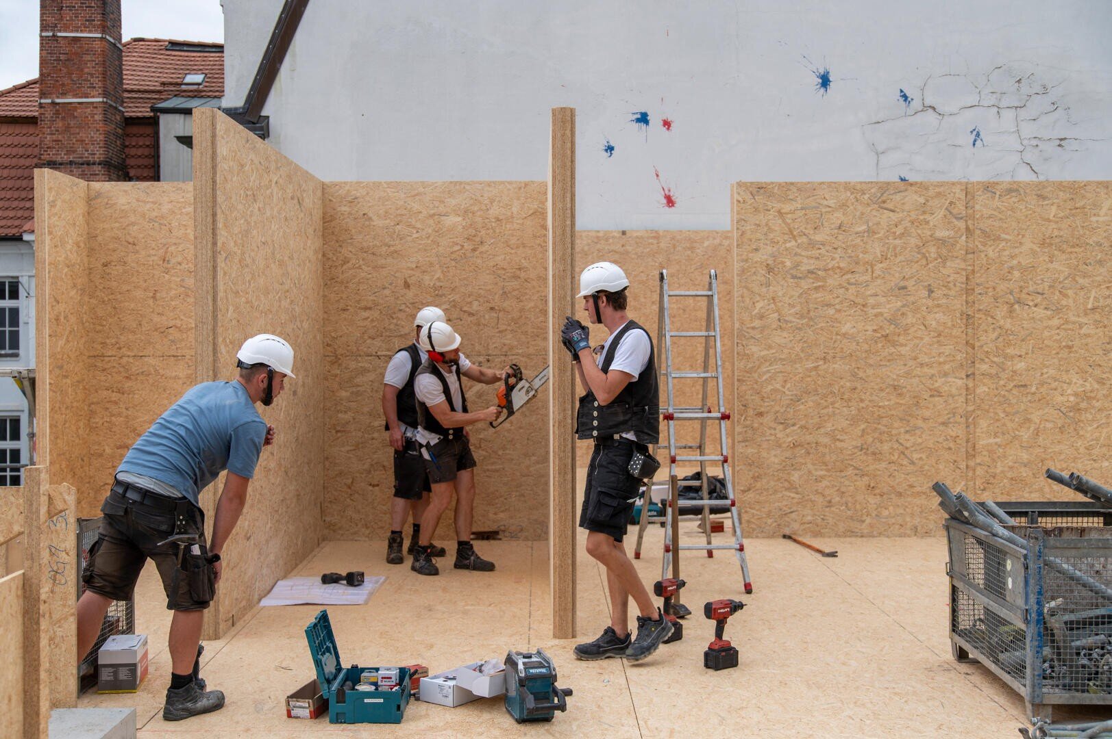 Three construction workers wearing helmets and safety equipment are assembling wooden panels on a building site. One is holding a beam upright, another is using a nail gun, and tools are scattered on the ground. A ladder leans against a wall.