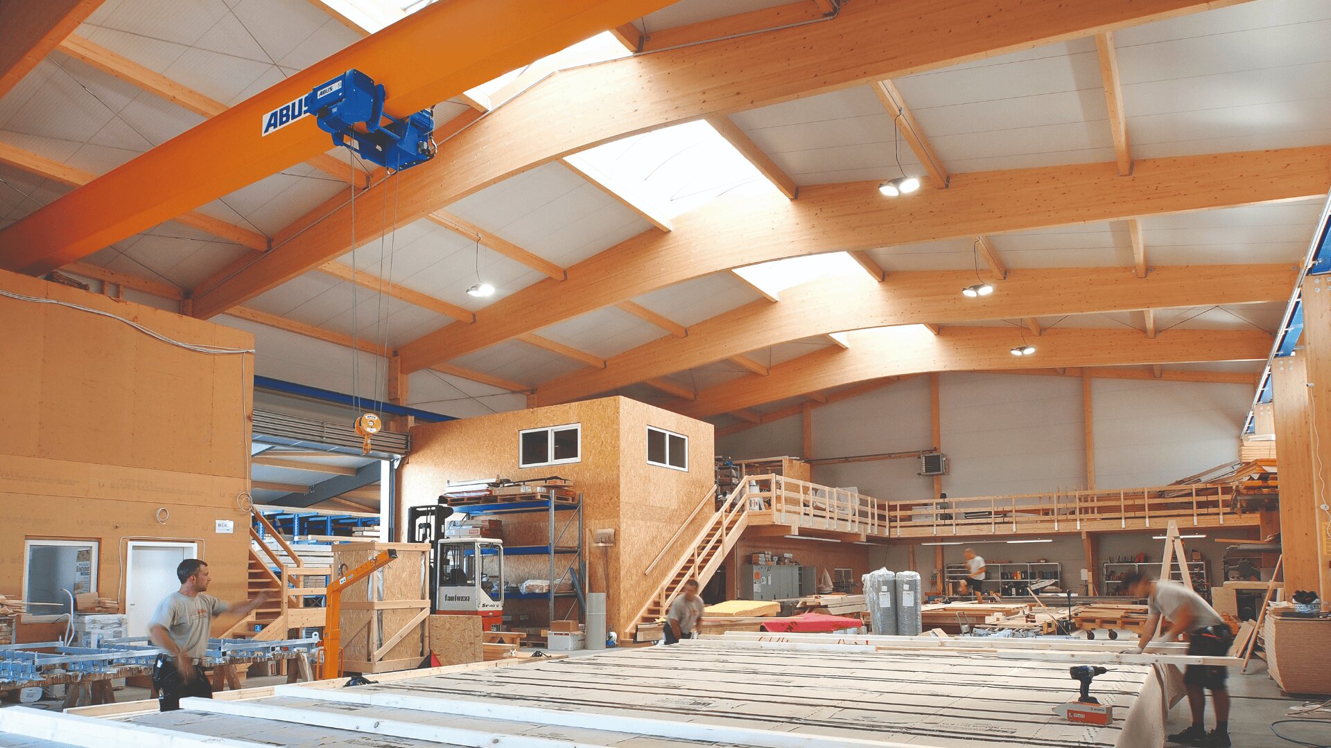 Innenraum einer geräumigen Holzwerkstatt mit hohen Decken, großen Holzbalken und Oberlichtern. Arbeiter montieren große Holzplatten. Im gesamten Arbeitsbereich sind verschiedene Werkzeuge und Geräte zu sehen.