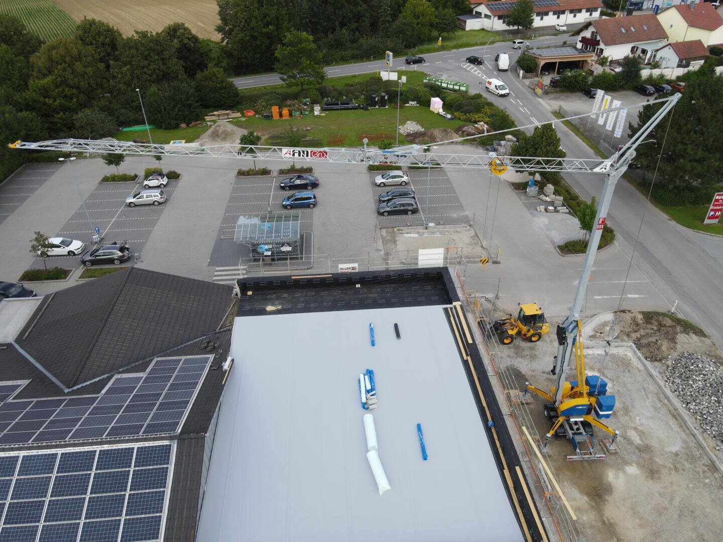 Luftaufnahme einer Baustelle mit einem großen Kran, geparkten Fahrzeugen, Sonnenkollektoren auf einem Dach und einem nahe gelegenen Parkplatz. Die umliegenden Straßen, Bäume und mehrere Gebäude sind ebenfalls zu sehen.