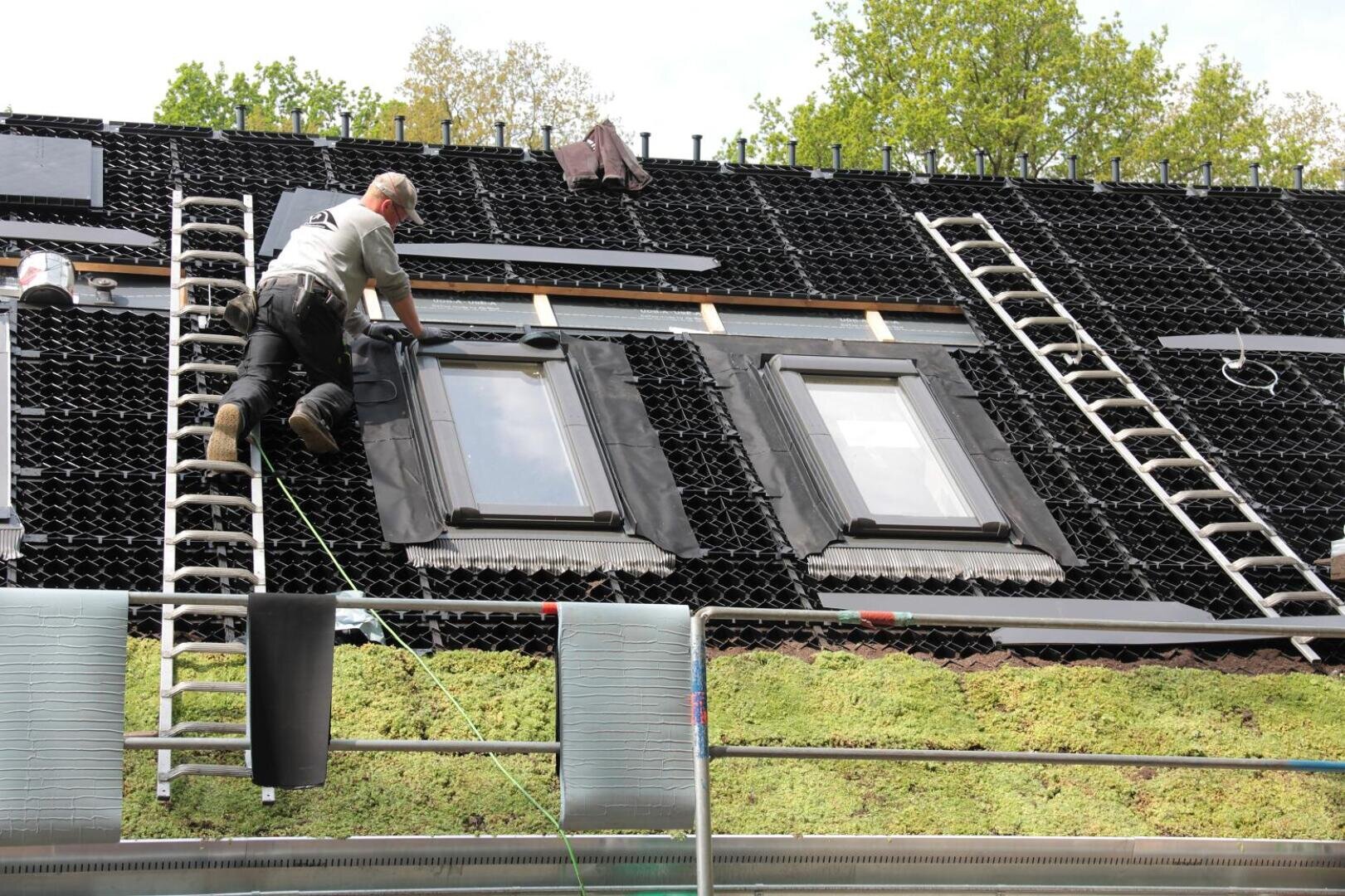 A worker installs roofing material around two skylight windows on a sloped roof partially covered with green vegetation. Ladders, tools, and roof materials are visible around him. Trees are in the background.