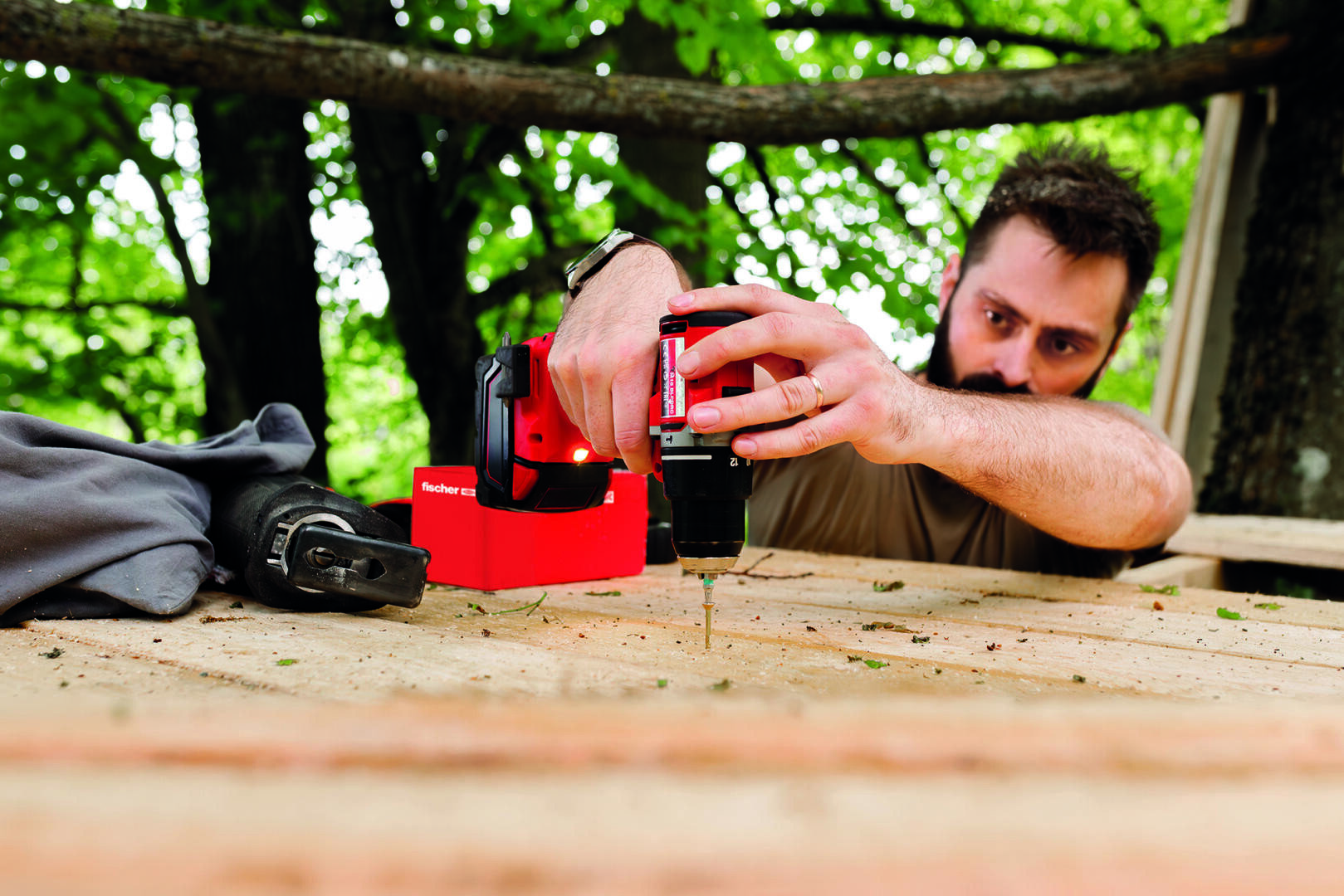 Ein Mann schraubt mit einer Bohrmaschine in eine Holzoberfläche im Freien, wobei im Hintergrund Bäume und Grünflächen zu sehen sind. Er konzentriert sich auf seine Aufgabe, und die Werkzeuge sind in der Nähe platziert.