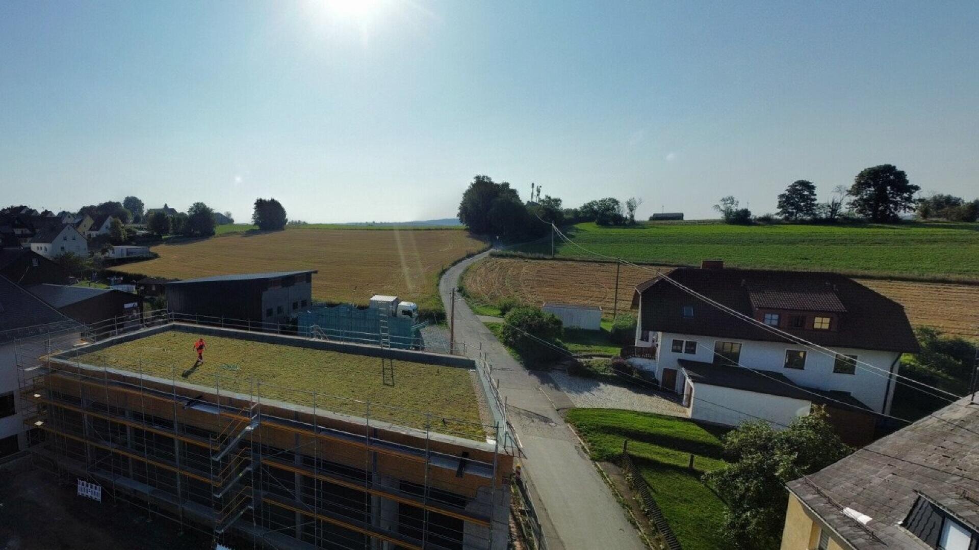 Aerial view of a rural area with fields, houses, and a road. One person stands on the grass-covered rooftop of a building under construction in the foreground. The sun is shining in a clear blue sky.