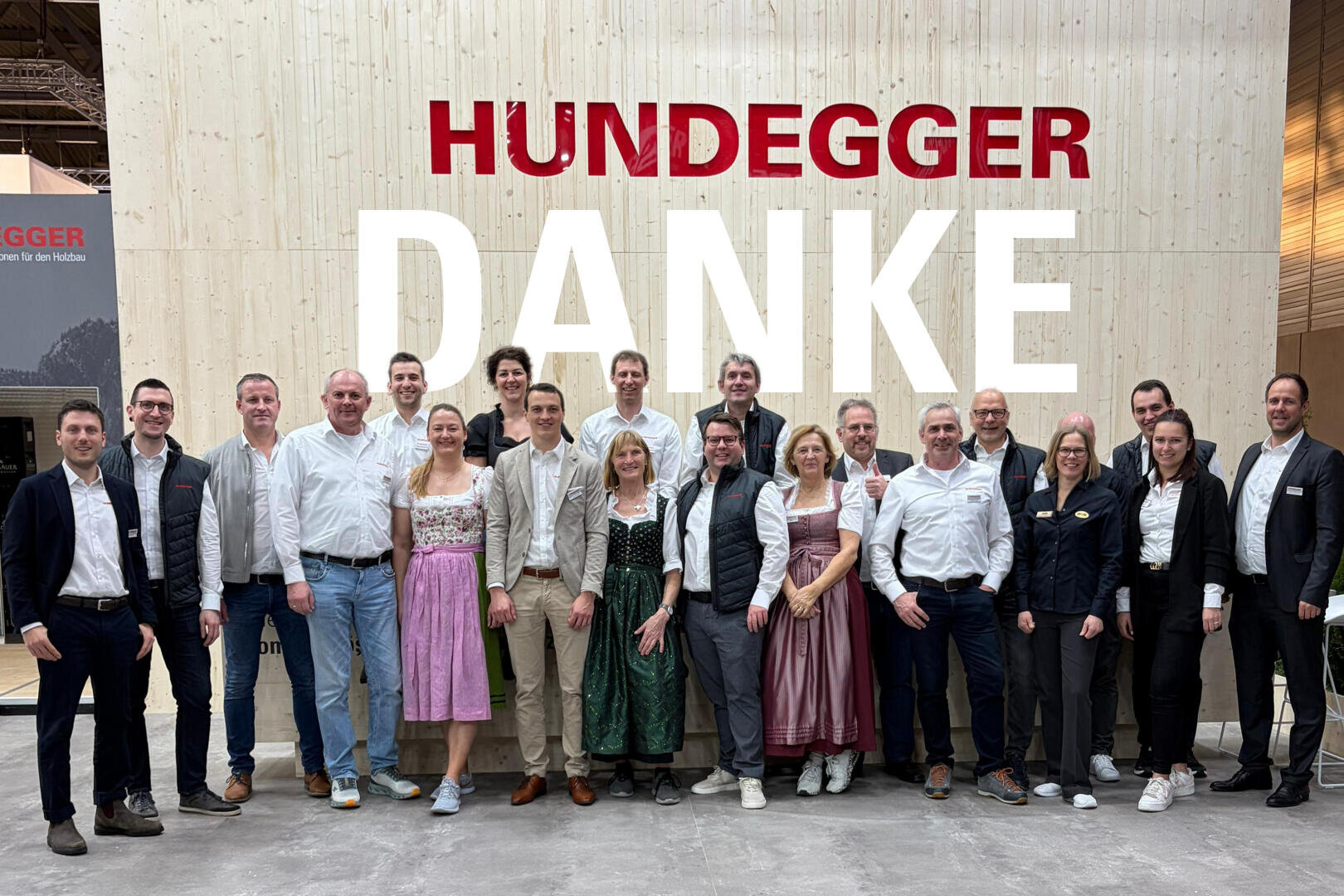 Eine Gruppe von Menschen steht lächelnd vor einer Holzwand mit der Aufschrift HUNDEGGER DANKE in großen Buchstaben; einige tragen traditionelle bayerische Kleidung, andere Geschäftskleidung.