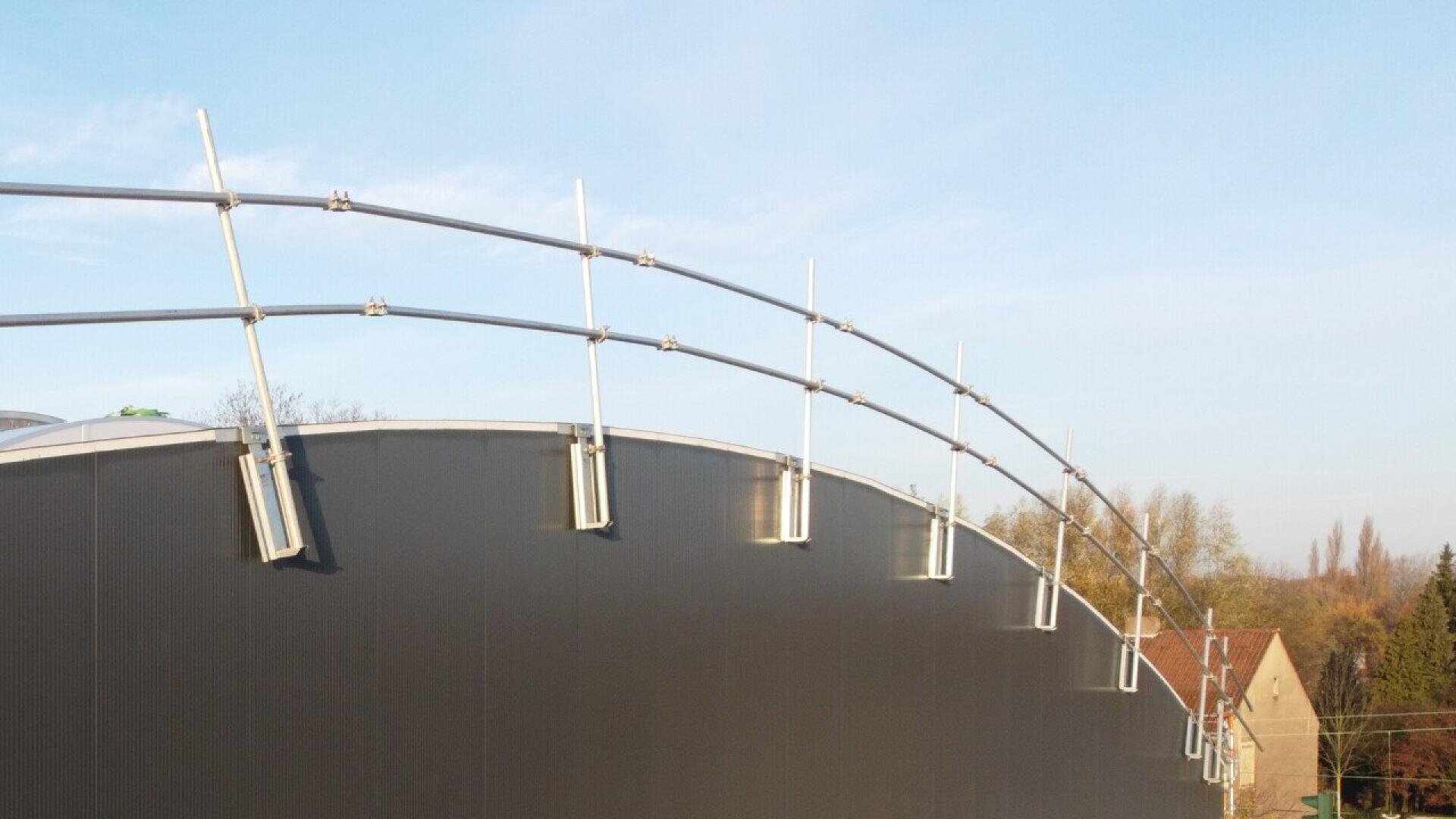 Curved metal guardrails are mounted along the edge of a large, dark-colored industrial roof. Trees and rooftops are visible in the background under a clear sky.