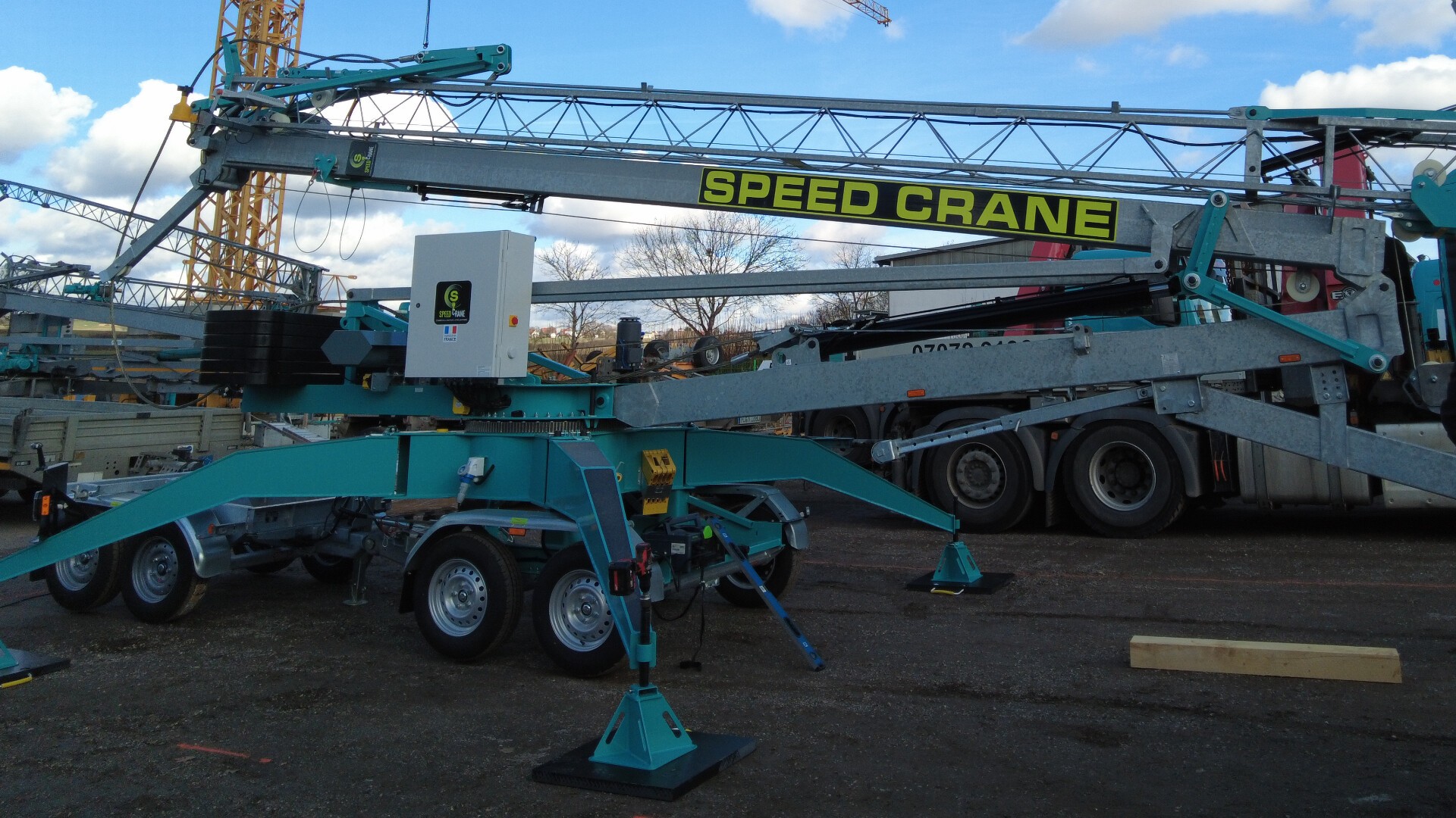 Ein großer Mobilkran mit der Aufschrift „SPEED CRANE“ steht mit ausgefahrenen Stützbeinen auf einer Baustelle. Im Hintergrund sind Geräte und ein LKW unter einem leicht bewölkten Himmel zu sehen.