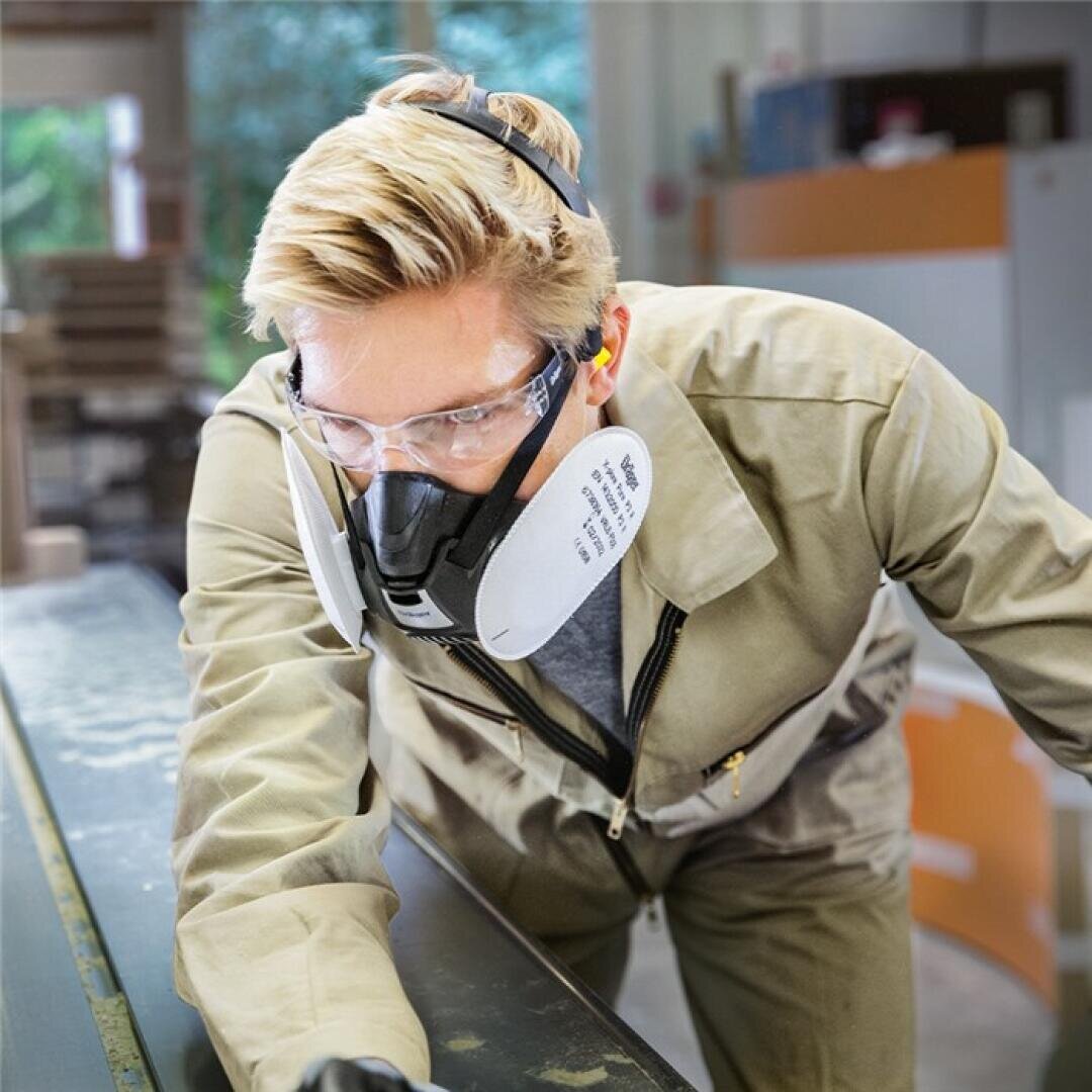 Eine Person, die eine Schutzbrille, eine Atemschutzmaske und einen beigen Overall trägt, arbeitet in einer Werkstatt und beugt sich über eine mit Staub oder Pulver bedeckte Fläche.