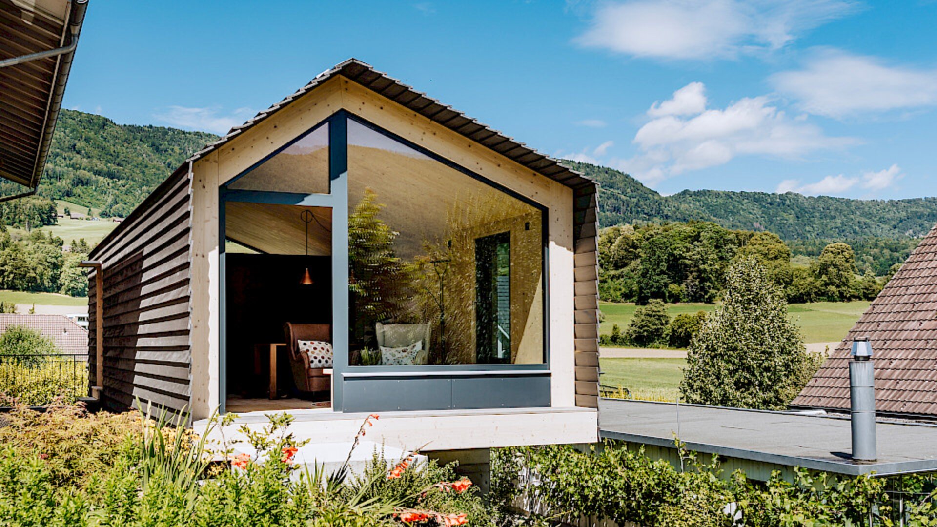 Eine moderne Holzhütte mit einem großen dreieckigen Glasfenster bietet unter einem hellen, teilweise bewölkten Himmel Aussicht auf üppige grüne Felder und Berge in der Ferne.