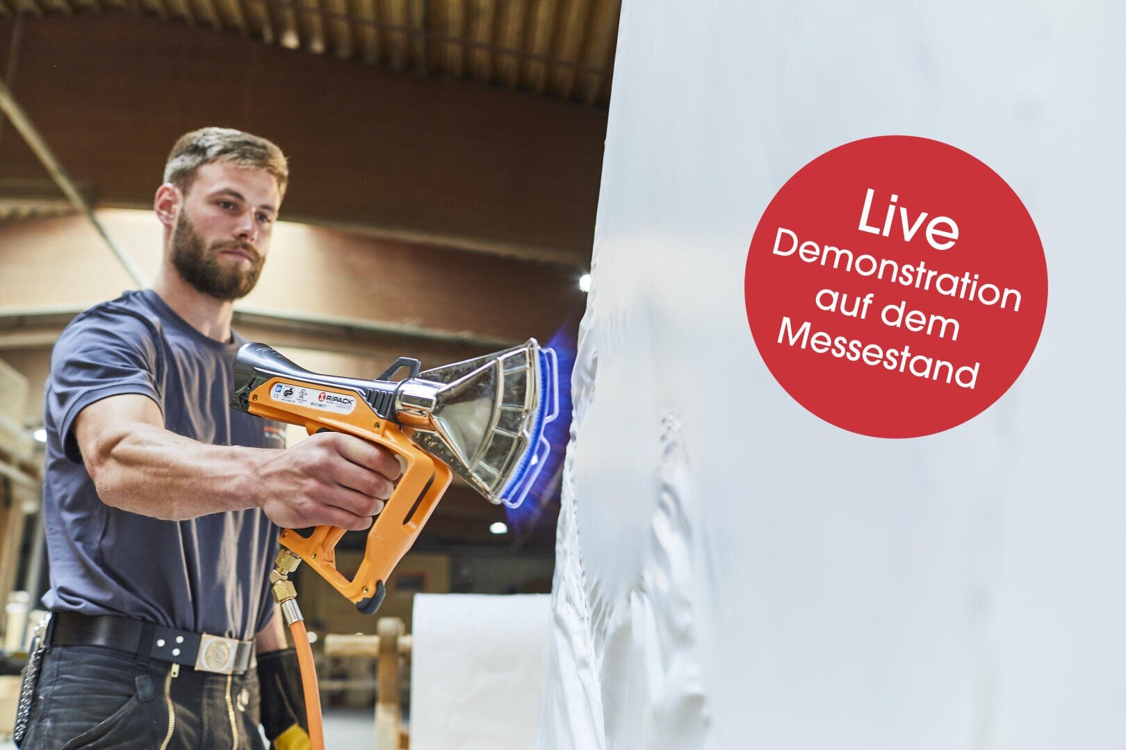Ein Mann benutzt eine Heißluftpistole auf einer großen weißen Fläche. Ein roter Kreis auf der rechten Seite zeigt den deutschen Text: Live-Demonstration auf dem Messestand. Der Schauplatz scheint ein Arbeitsraum oder eine Ausstellung zu sein.