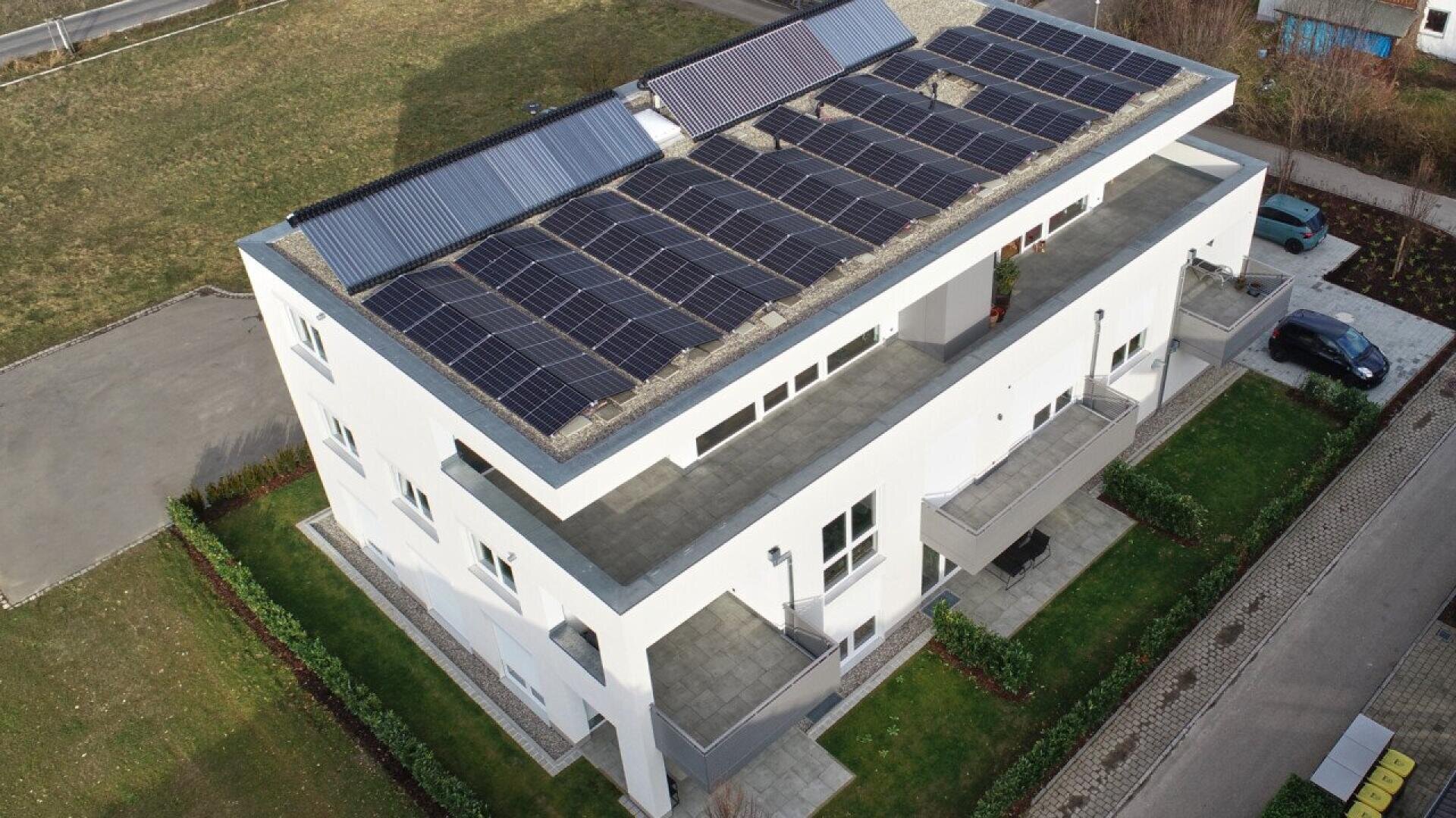 Aerial view of a modern white building with multiple balconies and solar panels installed on the flat roof, surrounded by green lawns and paved pathways.