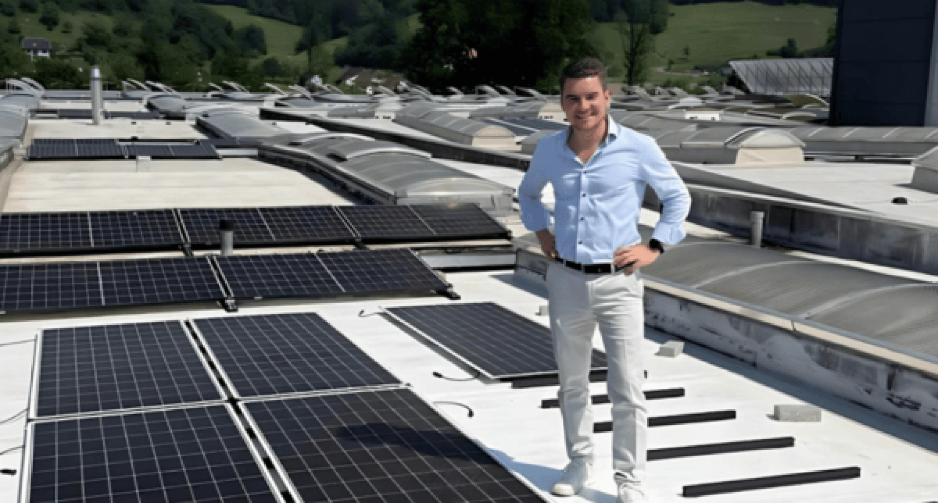 Ein Mann in hellblauem Hemd und weißer Hose steht auf einem Dach neben Reihen von Solarmodulen, im Hintergrund ist eine grüne, hügelige Landschaft zu sehen.