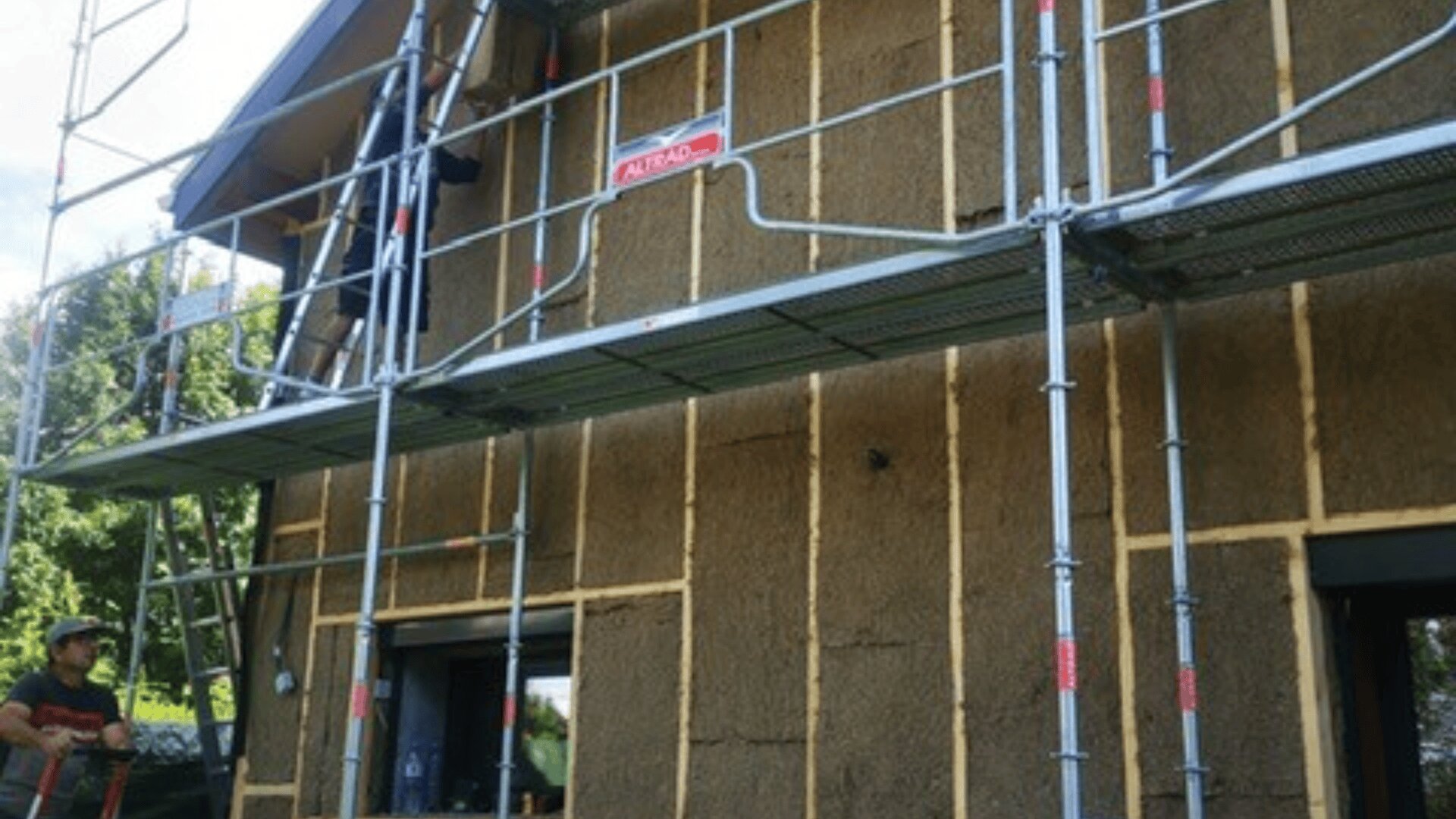 Eine Hausfassade im Bau mit sichtbarem Gerüst und Strohdämmung. Zwei Arbeiter sind anwesend, einer steht auf dem Boden und der andere auf dem Gerüst in der Nähe eines oberen Fensters.