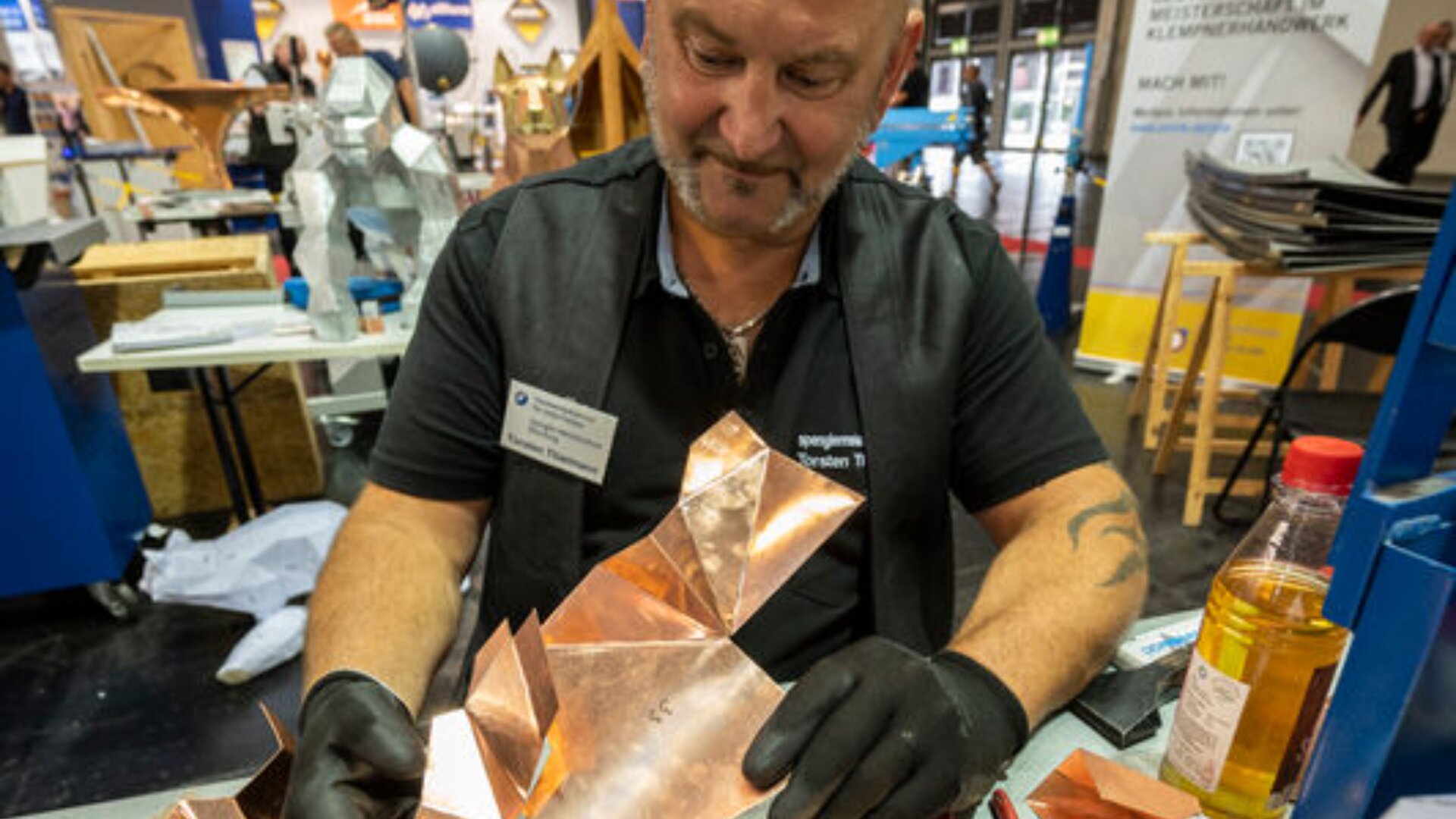 Ein Mann mit Handschuhen faltet und formt ein Kupferblech an einem Werkstatttisch, umgeben von Metallbearbeitungswerkzeugen und -materialien.