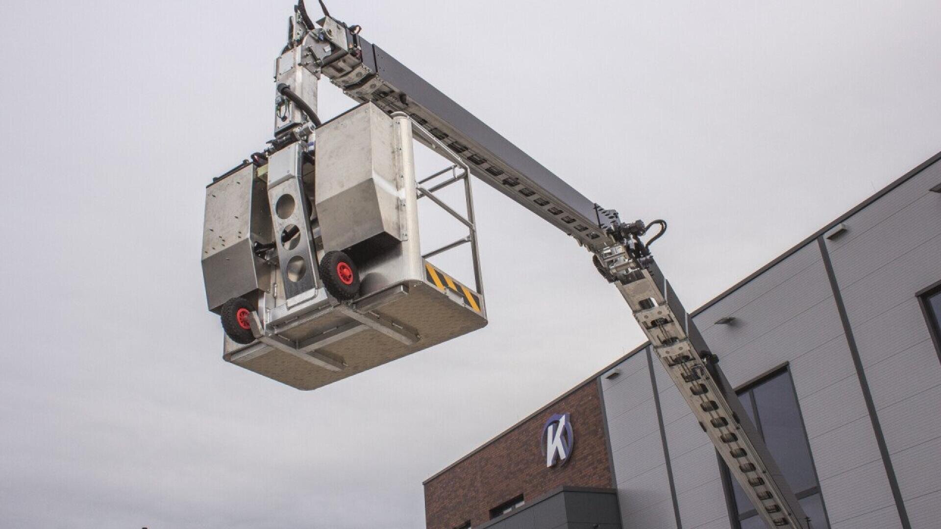 A close-up of a large industrial lift platform raised high beside a modern building with a gray facade and a brick section displaying a white K logo under a cloudy sky.