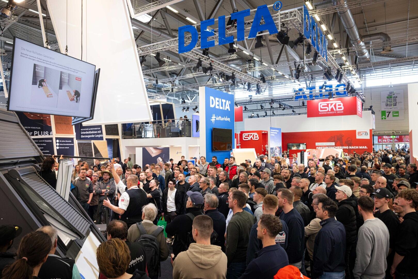 Eine große Menschenmenge versammelt sich an einem Messestand unter einem blauen "DELTA"-Schild und verfolgt eine Präsentation. Zahlreiche Banner, Displays und Produktexponate füllen die helle, geschäftige Ausstellungshalle.