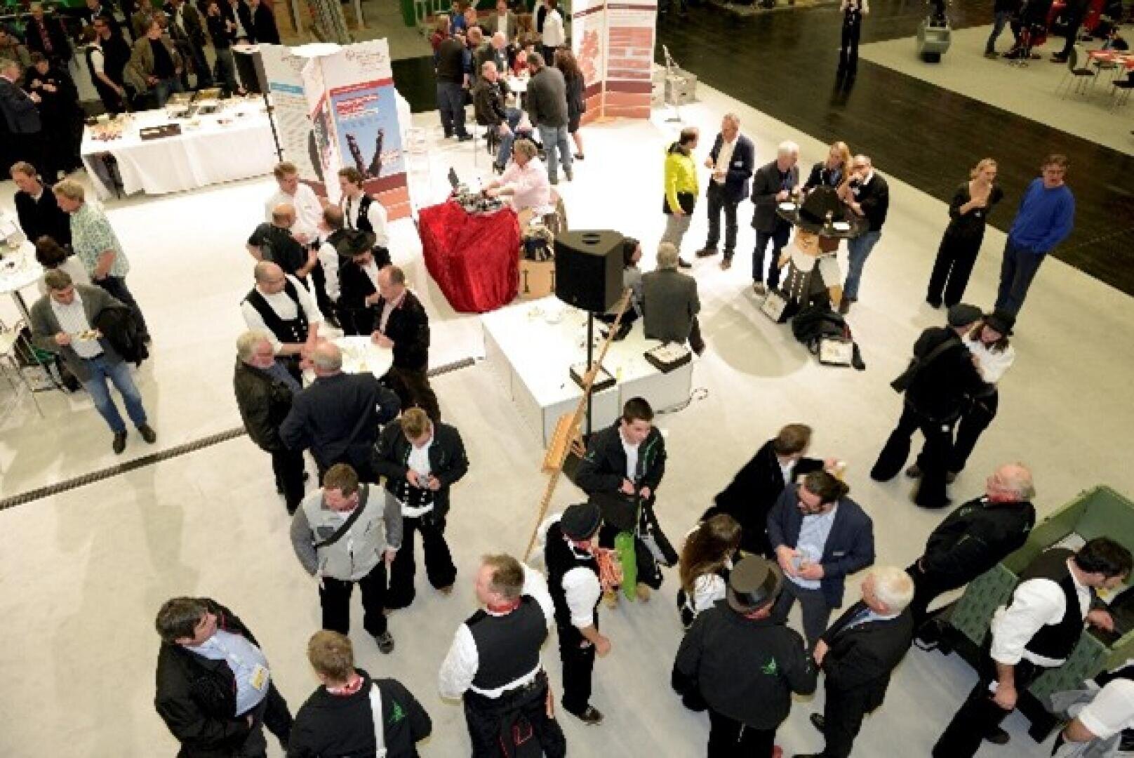 Eine überfüllte Veranstaltung in einem Gebäude, bei der Menschen in Gruppen stehen und sich unterhalten. Tische mit Speisen und Getränken sind zu sehen, und die Teilnehmer tragen Geschäfts- und Freizeitkleidung. Einige Personen scheinen Kontakte zu knüpfen oder sich zu unterhalten.