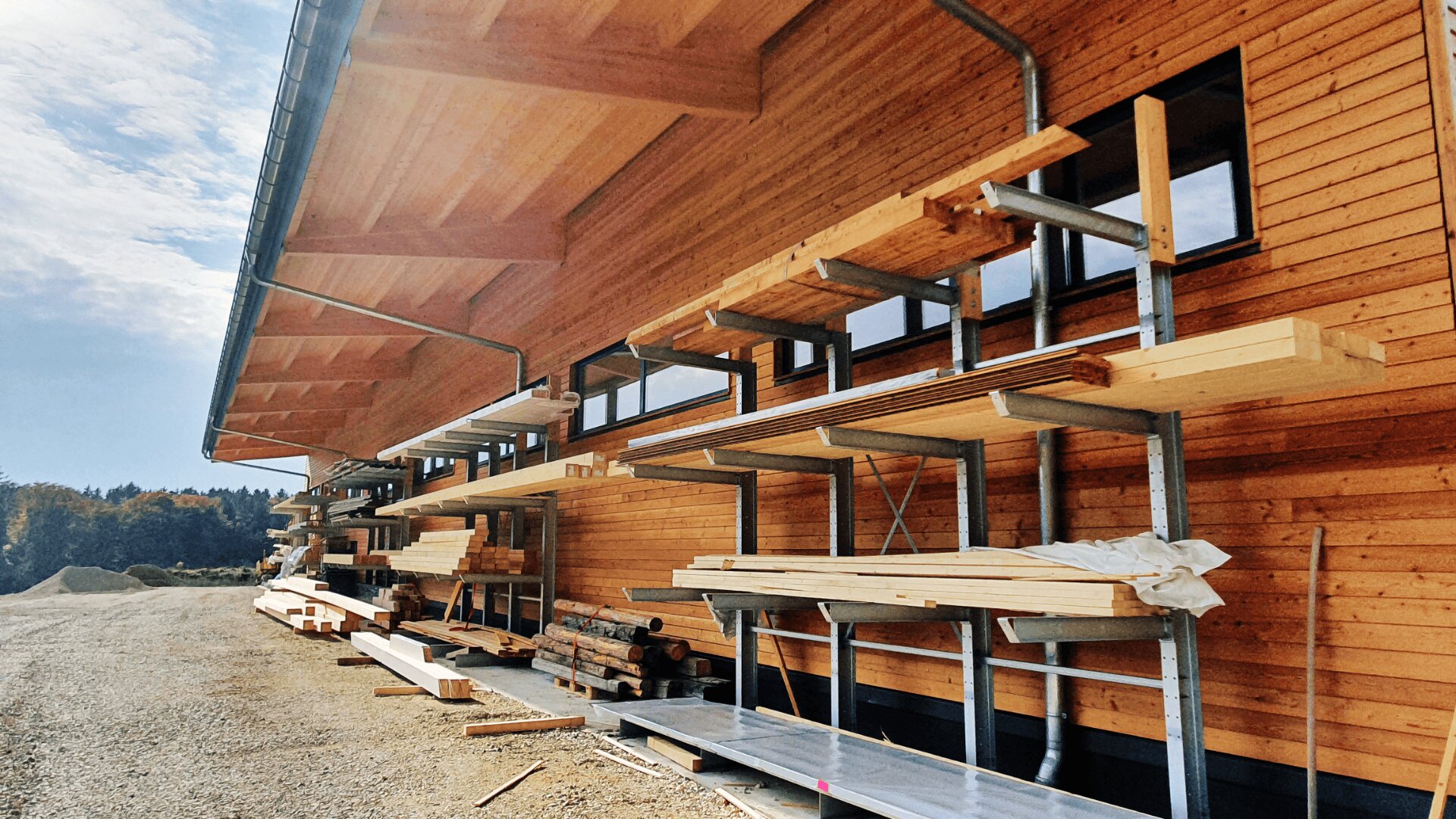 Stapel aus Holzbrettern und -balken sind ordentlich auf Metallgestellen gelagert, die an der Außenwand eines großen Holzgebäudes unter einem schrägen Dach befestigt sind, neben dem sich eine Kiesbereich befindet.
