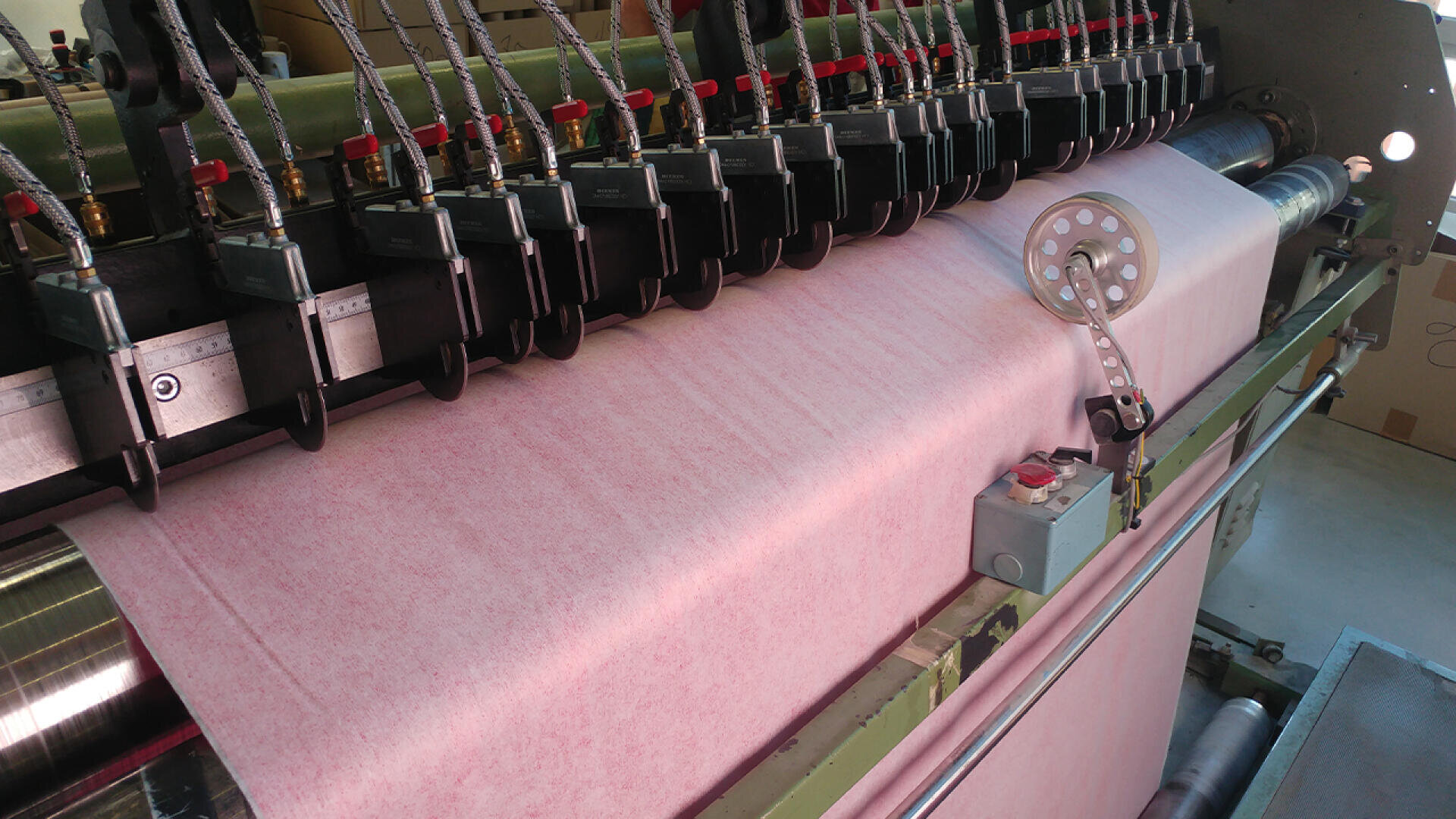 Eine Nahaufnahme einer Industriemaschine, die eine Rolle hellrosa Stoff verarbeitet, mit mehreren schwarzen und roten Komponenten und einem seitlich sichtbaren Bedienfeld.