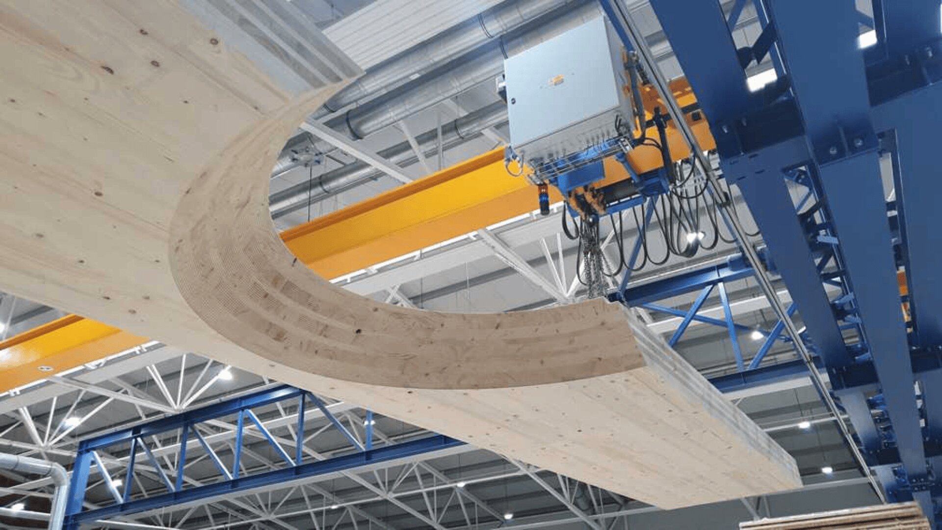 Ein großer gebogener Holzbalken wird von einem gelben Brückenkran in einer Industrieanlage mit Metallbalken und heller Beleuchtung angehoben.