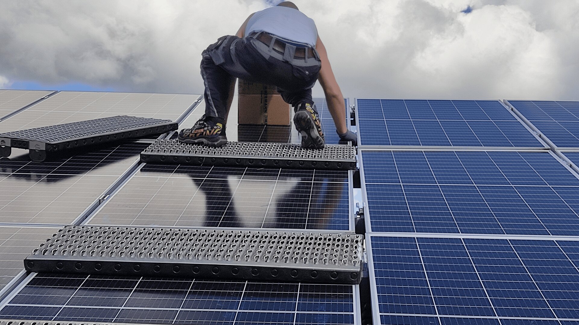 A person in work boots and gloves is installing or maintaining solar panels on a rooftop, standing on metal steps placed across the panels under a cloudy sky.
