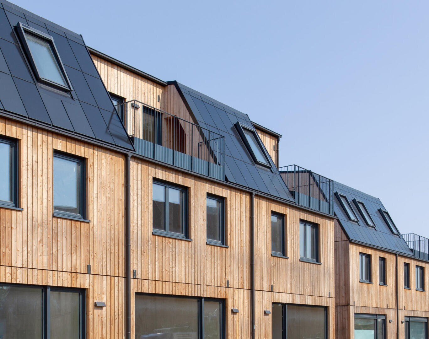 Eine Reihe moderner Stadthäuser mit Holzfassaden und schwarzen Metalldächern mit vielen Fenstern und Oberlichtern vor einem klaren blauen Himmel.