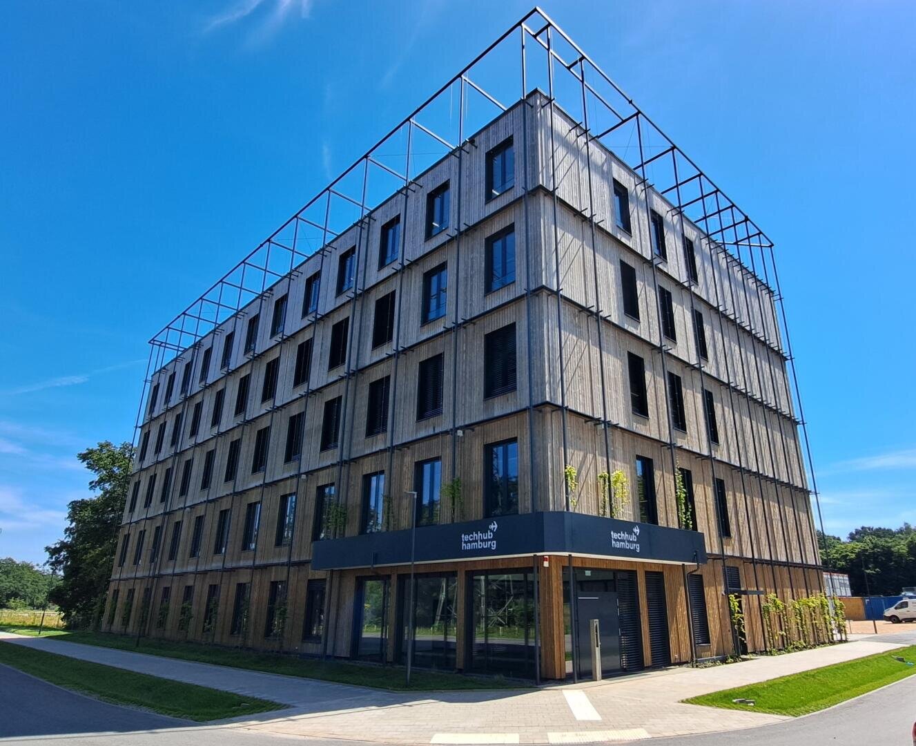 A modern, four-storey office building with large windows and a wood-panelled façade on which the logo of Ingolstadt University of Applied Sciences can be seen against a bright blue sky.