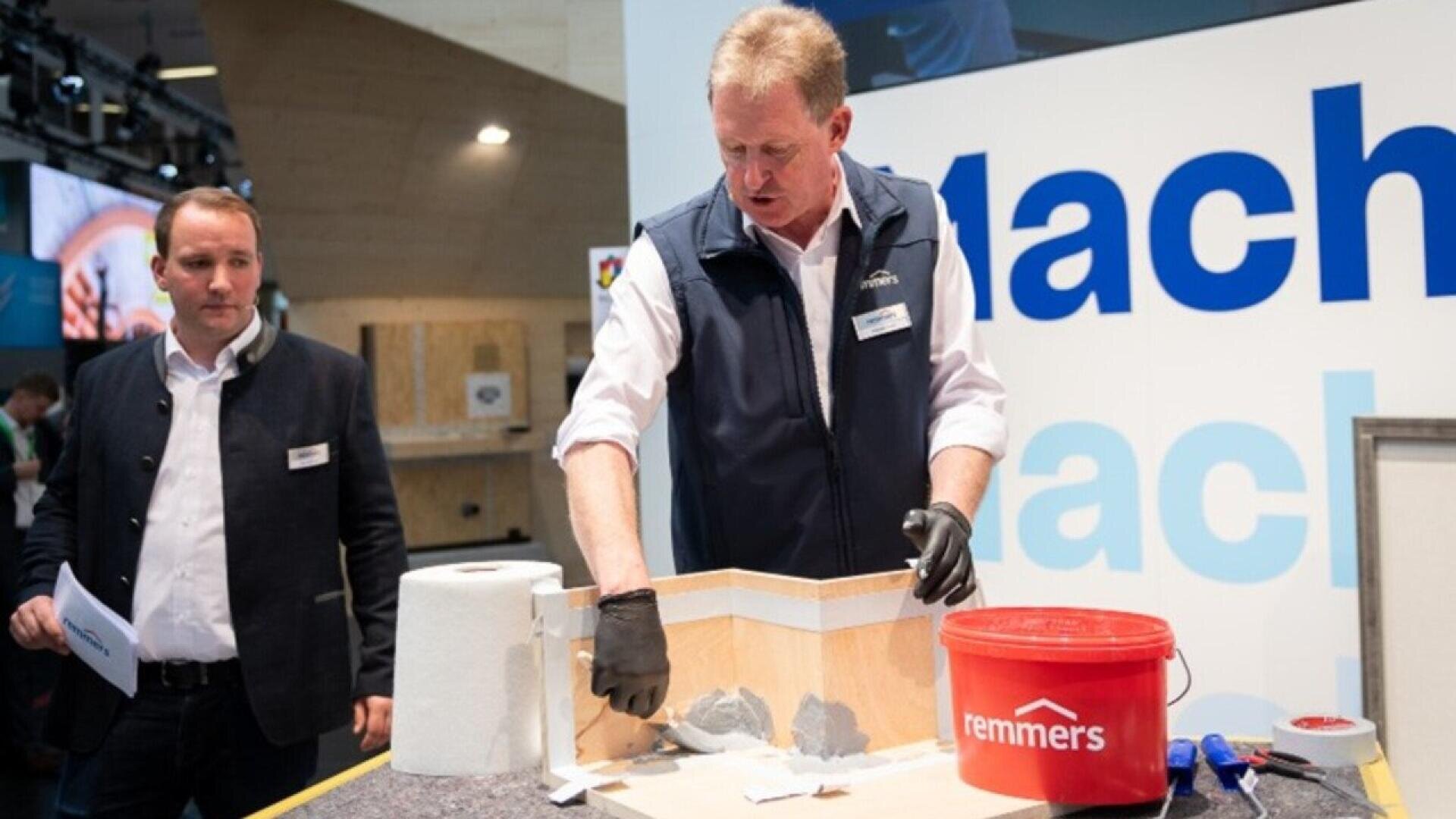 A man wearing gloves demonstrates applying a substance to a model structure, while another man stands nearby. There is a red Remmers bucket, paper towels, and tools on the counter. A large sign is visible in the background.