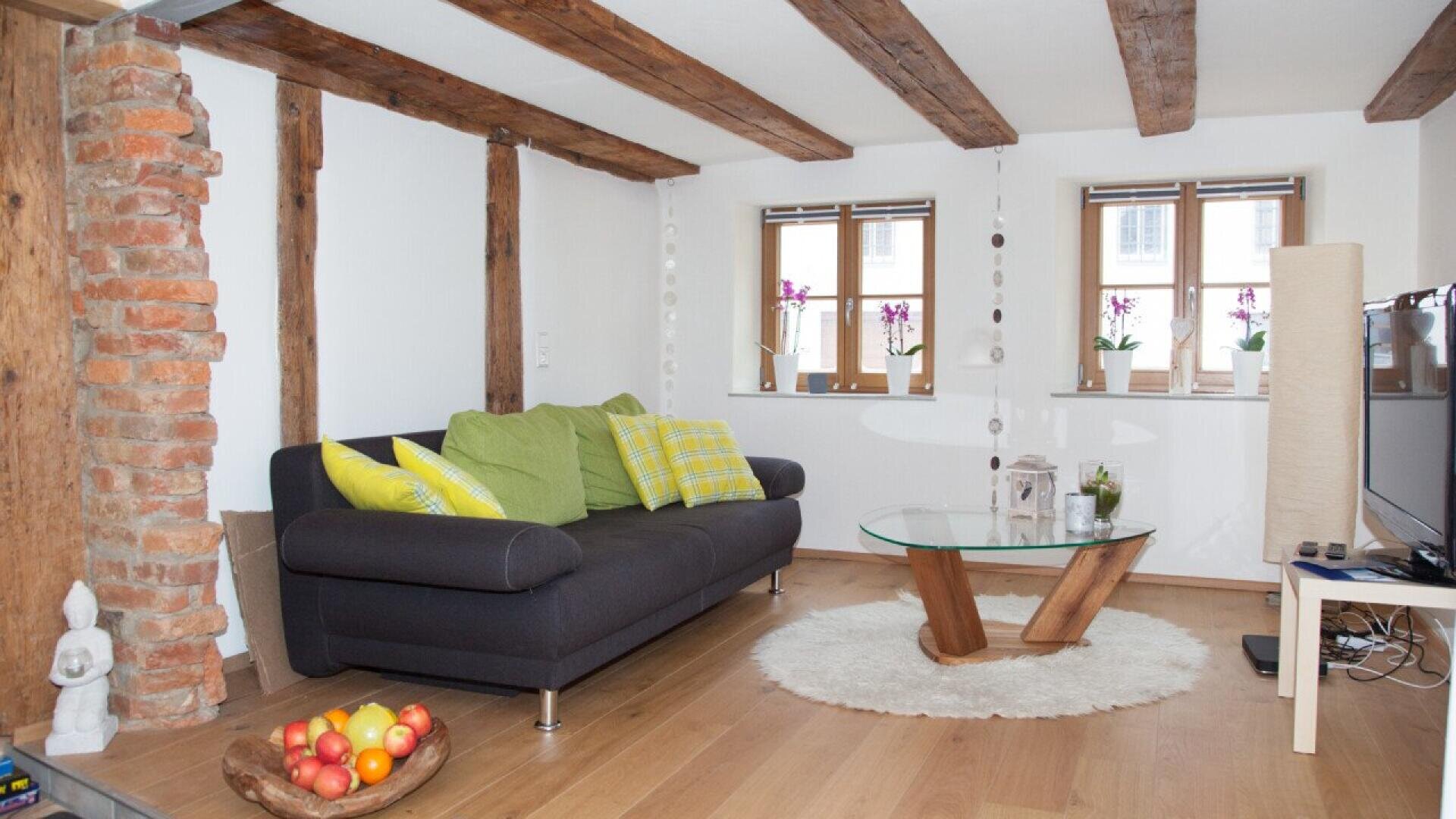 A cozy living room with exposed wooden beams, a dark gray sofa with green and yellow pillows, a glass coffee table, a shaggy white rug, potted plants on the windowsill, and a bowl of fruit on the wooden floor.