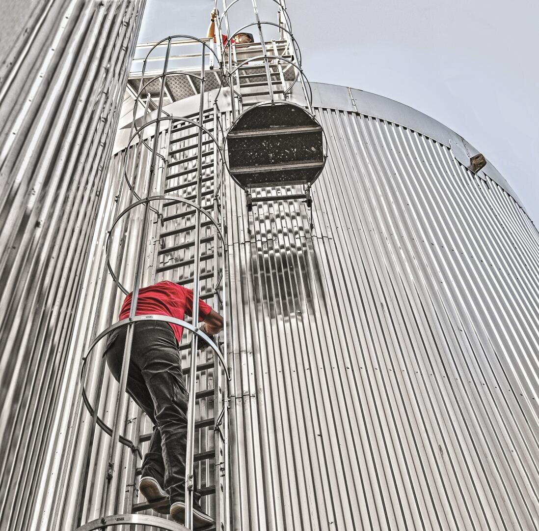 Eine Person, die ein rotes Hemd und eine dunkle Hose trägt, klettert auf eine mit einem Käfig versehene Metallleiter, die an der Seite eines großen, silbernen, gewellten Industriesilos befestigt ist, neben der eine weitere Leiter und ein weiteres Silo sichtbar sind.
