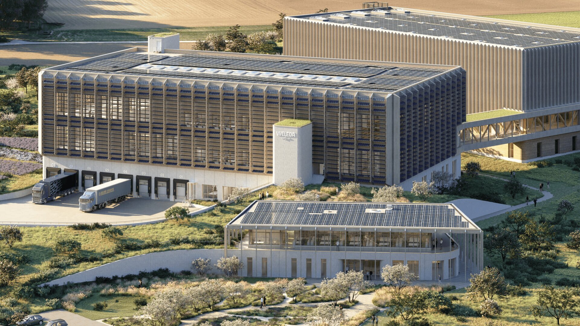 Eine moderne Industrieanlage mit Solarmodulen auf den Dächern, umgeben von Grün. LKWs parken an Laderampen, im Vordergrund steht ein kleineres Bürogebäude. Die Landschaft ist ländlich, im Hintergrund sind Felder zu sehen.