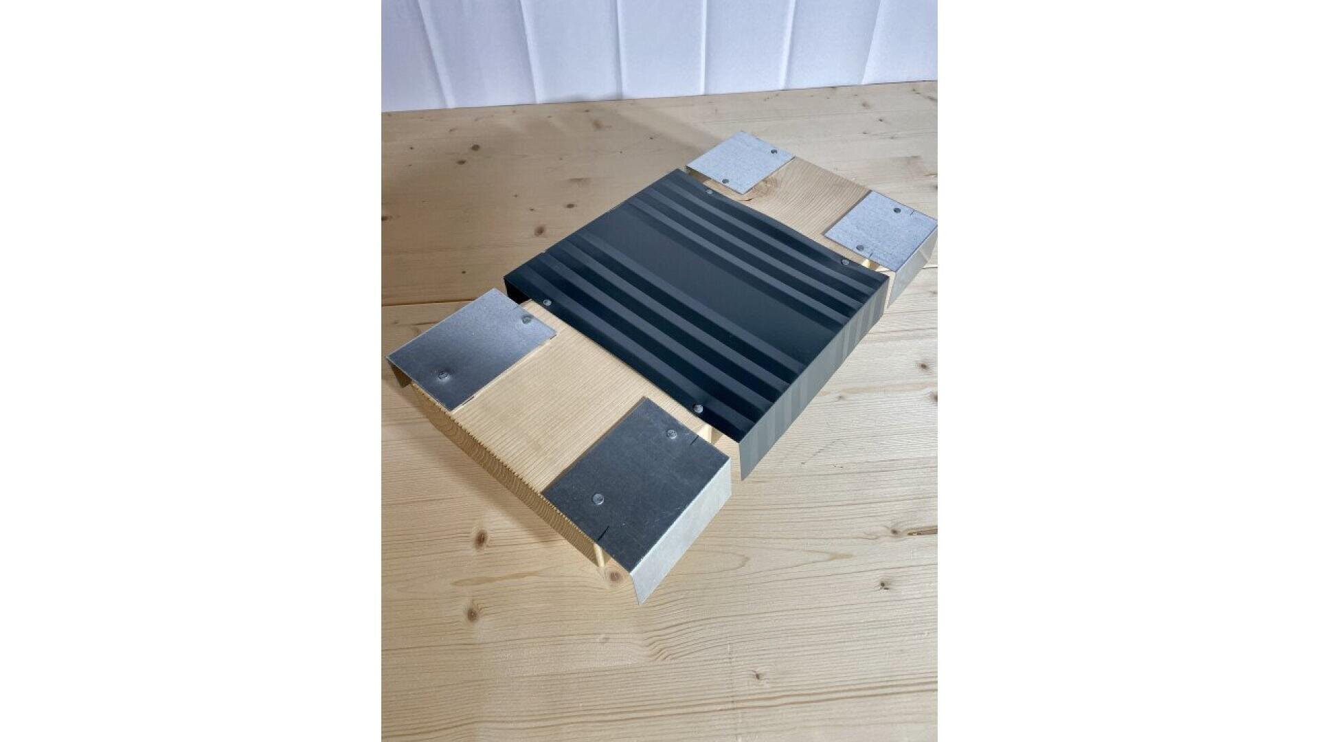 A wooden platform with four metal corner brackets and a central black grooved panel sits on a light wood surface against a white background.
