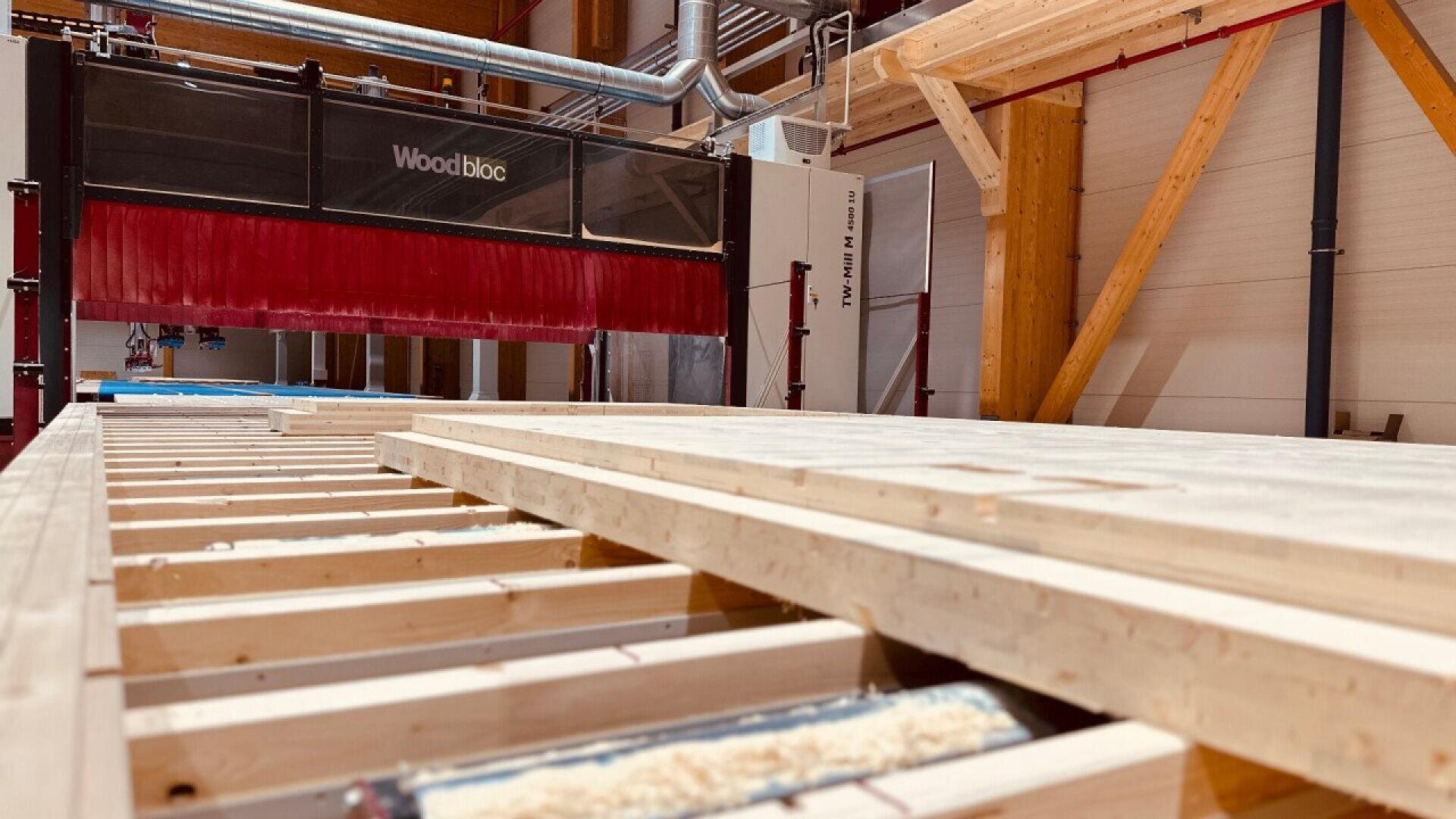 A large industrial woodworking machine labeled Woodbloc processes wooden beams inside a modern, well-lit facility with exposed wooden supports and red safety barriers.