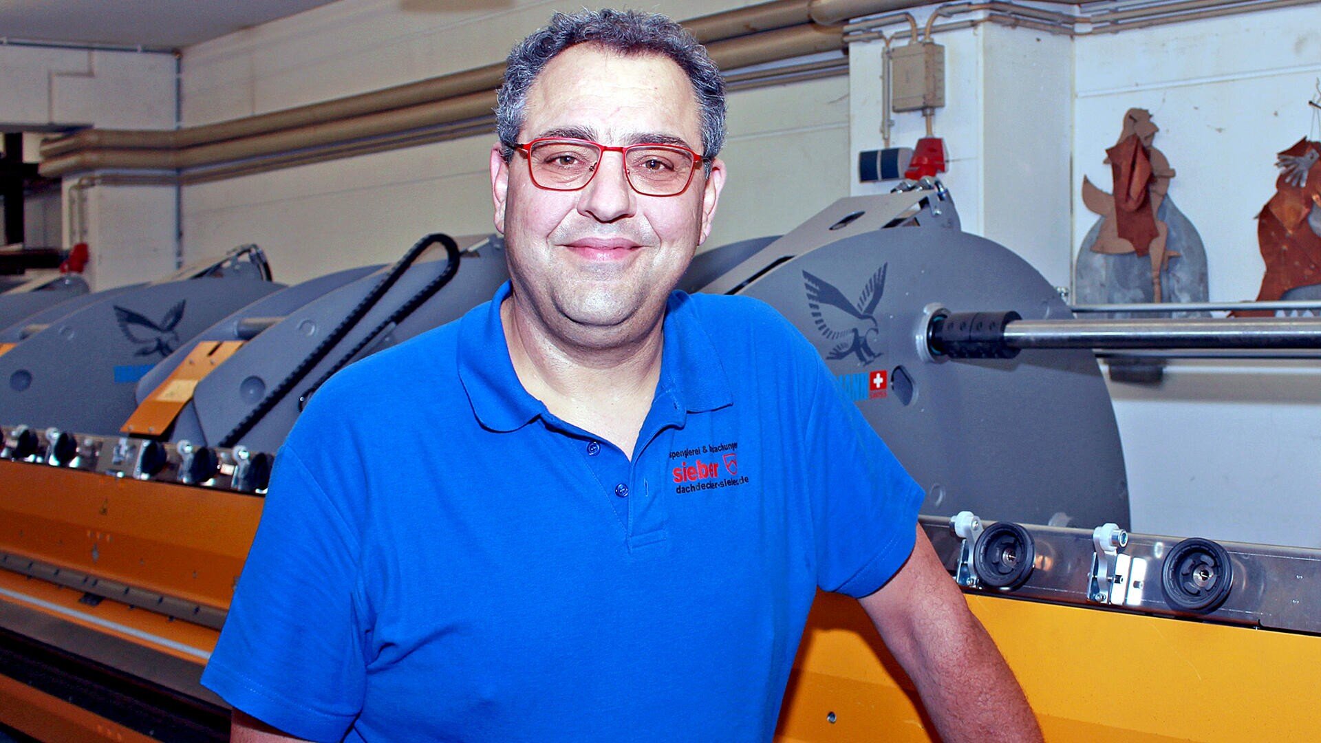 A man wearing glasses and a blue polo shirt stands smiling in front of industrial printing equipment inside a workshop.