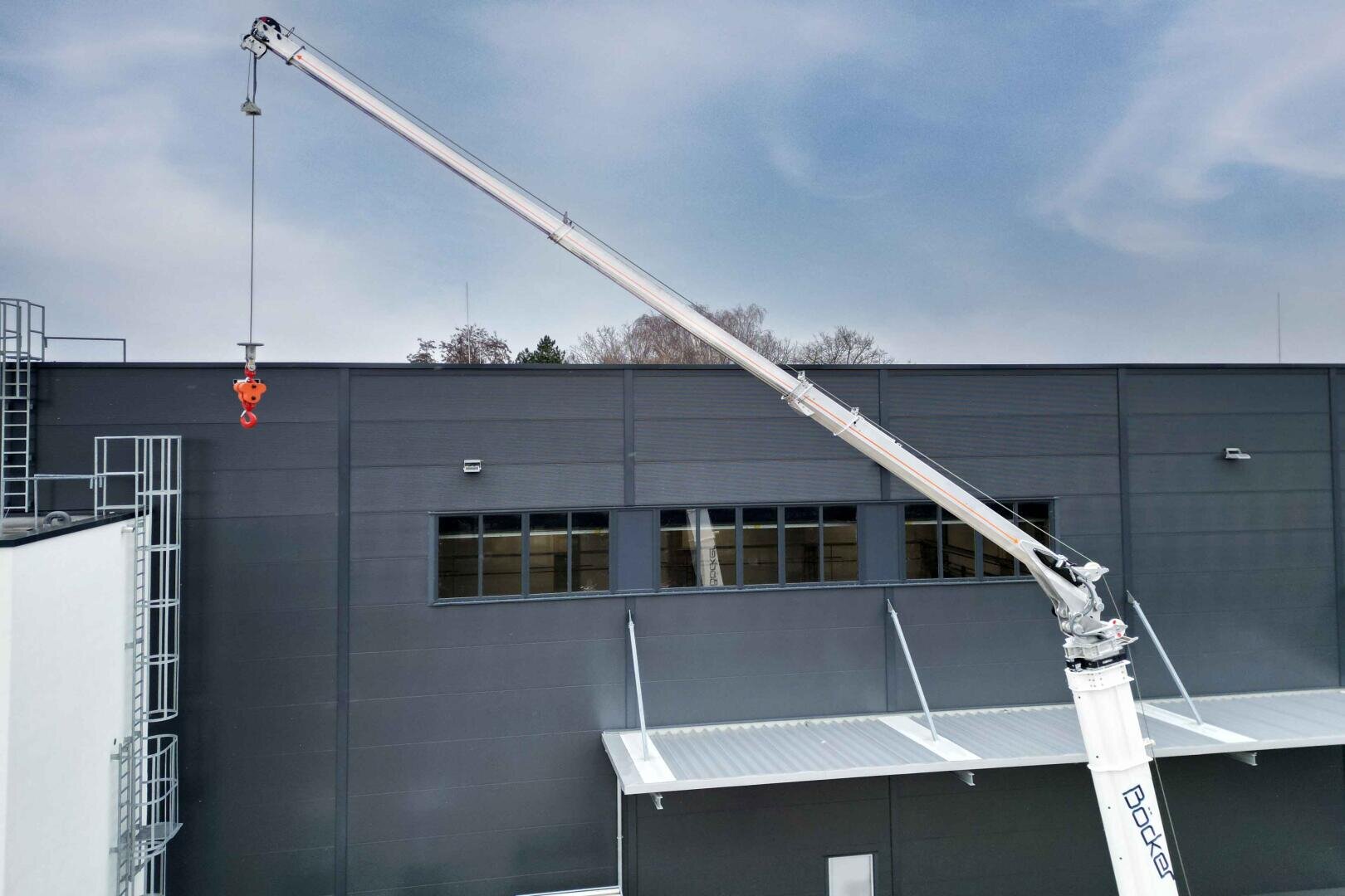 A large telescopic crane with a silver boom is extended over a modern industrial building and dangles with its red hook in front of high, dark gray walls and windows under a cloudy sky.