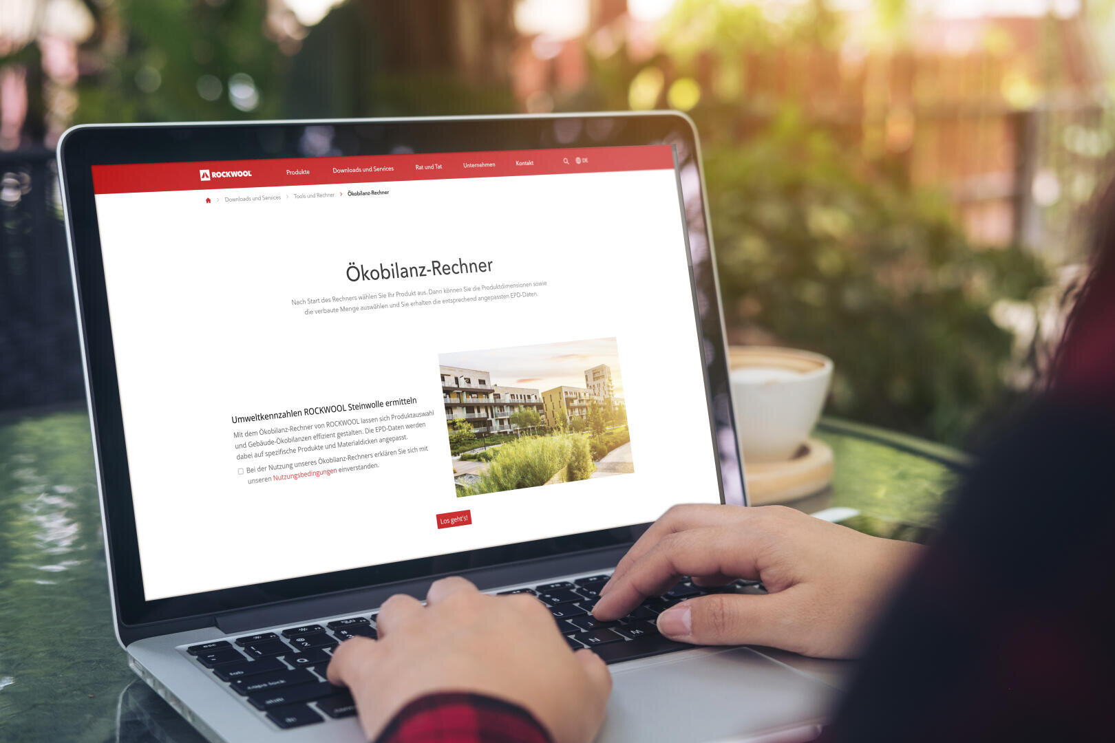 Eine Person tippt auf einem Laptop, der eine deutsche Webseite mit dem Titel Ökobilanz-Rechner und einem Bild moderner Gebäude anzeigt; im Hintergrund sind eine Kaffeetasse und Grünpflanzen zu sehen.