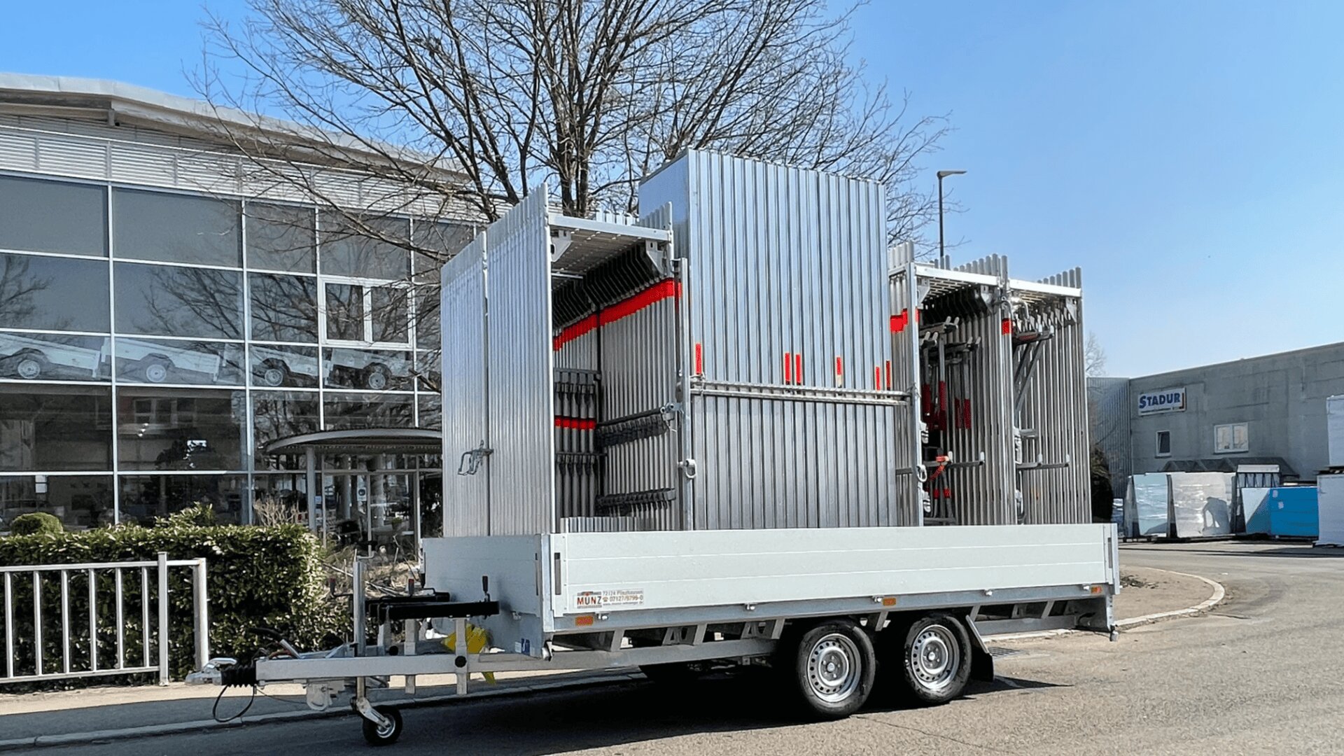 An einem sonnigen Tag wird eine große Metallkonstruktion mit vertikalen Paneelen und roten Akzenten auf einen weißen Anhänger geladen, der in der Nähe eines modernen Glasgebäudes auf einer Straße geparkt ist.