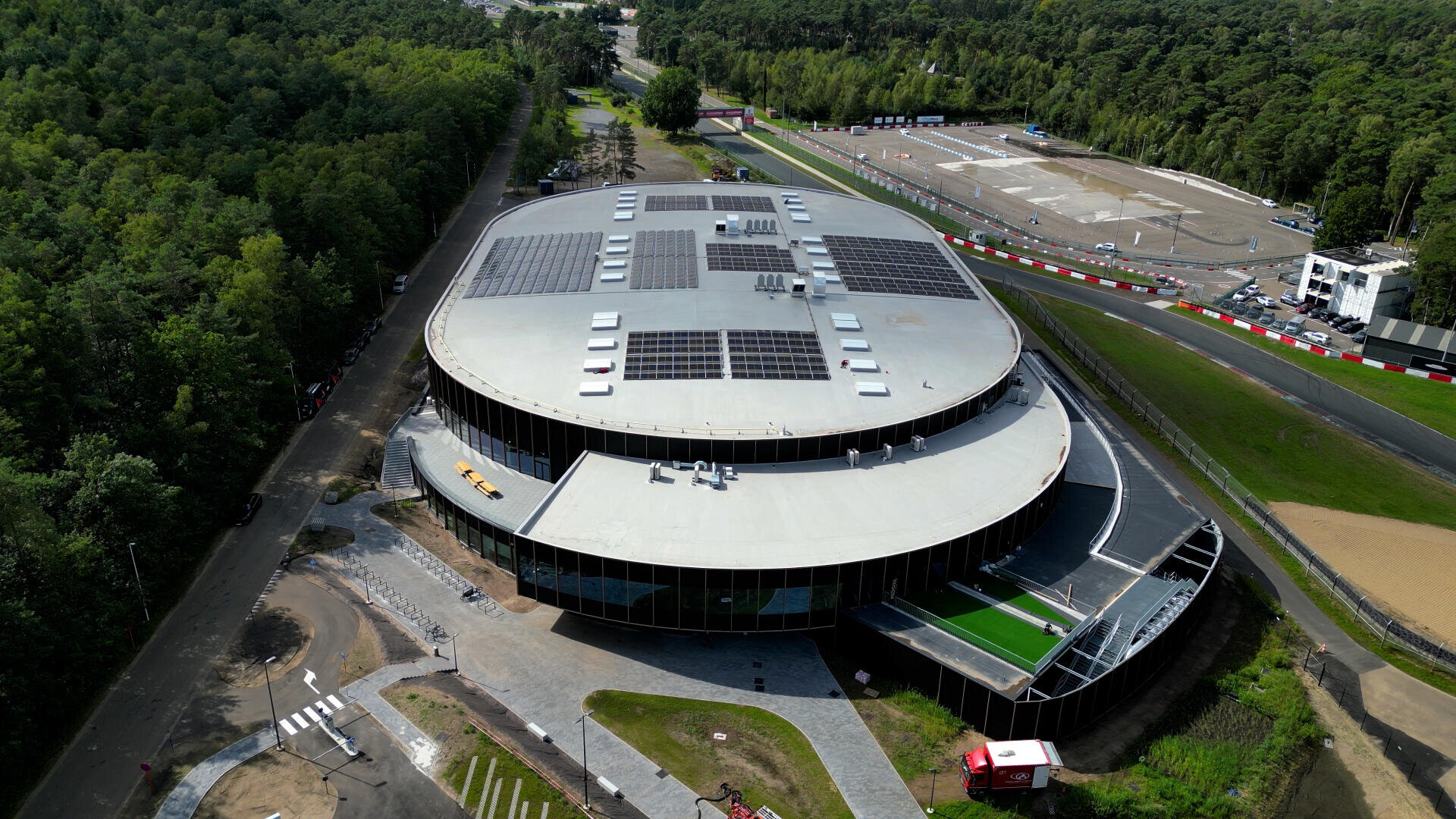 Luftaufnahme eines großen, modernen runden Gebäudes mit Sonnenkollektoren auf dem Dach, umgeben von Bäumen, Straßen und einer nahe gelegenen Rennstrecke mit rot-weißen Absperrungen.