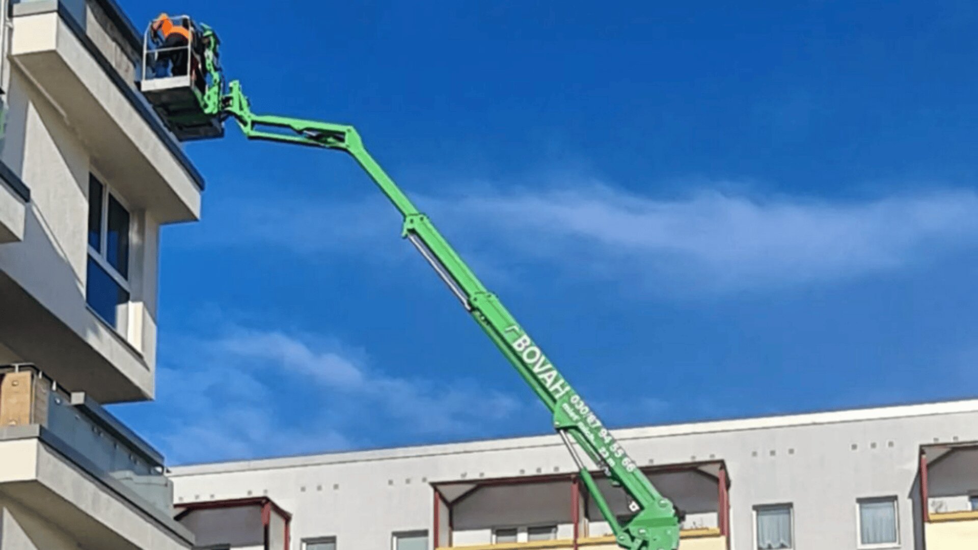 Ein Arbeiter in einer orangefarbenen Weste steht in einer hochgeklappten grünen Hebebühne und streckt sich vor einem klaren blauen Himmel bis zum Balkon eines hohen Gebäudes.