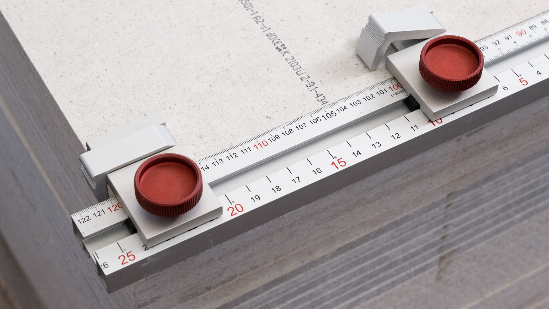 A close-up of a precision measuring device with red dials and white clamps on a metal ruler, set on a flat, light-colored surface marked with numbers and lines for measurement.
