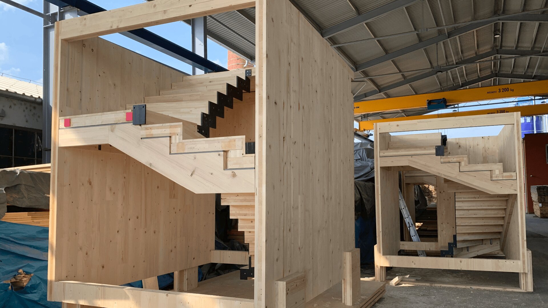In einer geräumigen Industriehalle mit freiliegenden Balken und teilweise offenem Dach entstehen derzeit zwei große Holztreppenkonstruktionen. Die Treppen bestehen aus hellem Holz mit schwarzen Metallkonsolen.