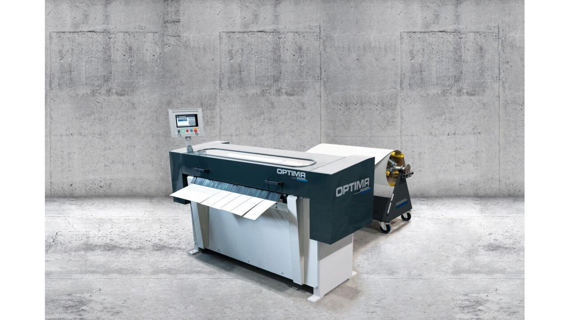 A large industrial machine labeled OPTIMA with a digital control panel and paper sheets being processed, set against a concrete wall in a factory or warehouse environment.
