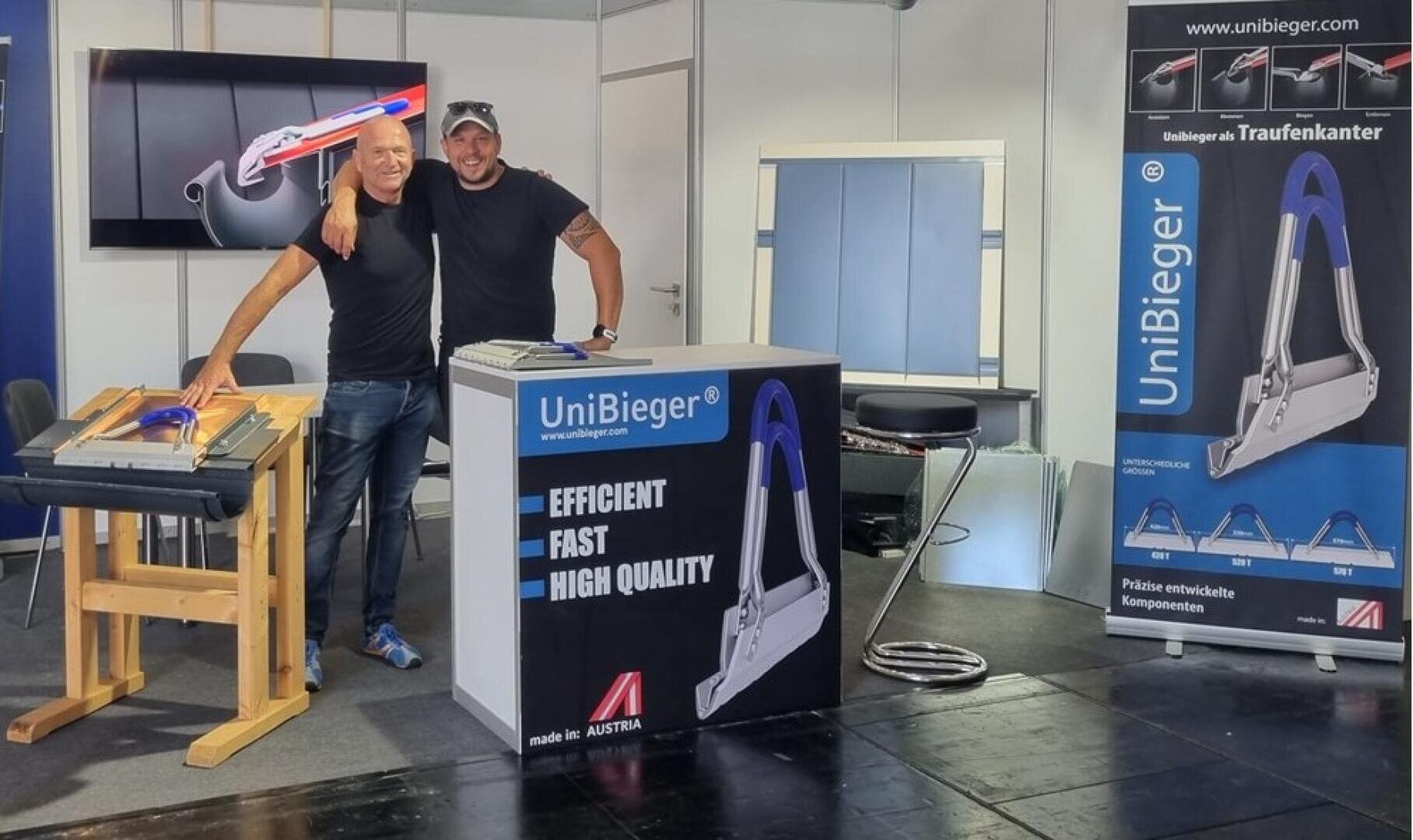 Zwei lächelnde Männer stehen zusammen an einem Messestand, der UniBieger-Metallbiegewerkzeuge ausstellt. Im Hintergrund sind Marketingbanner und Produktmuster zu sehen, die Effizienz, Geschwindigkeit und Qualität betonen.