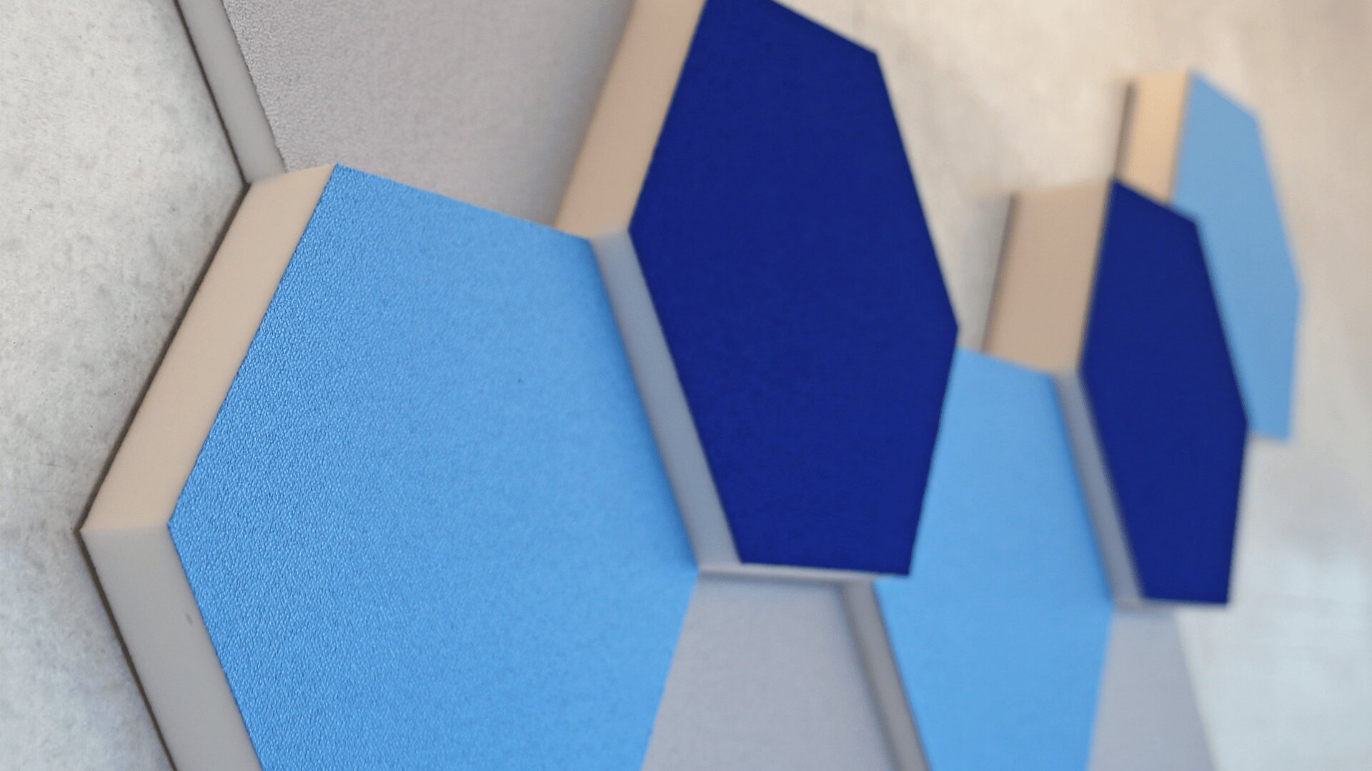 Sechseckige Akustikplatten in Blau- und Grautönen sind an einer hellen Wand angebracht und erzeugen ein geometrisches, modernes Design.