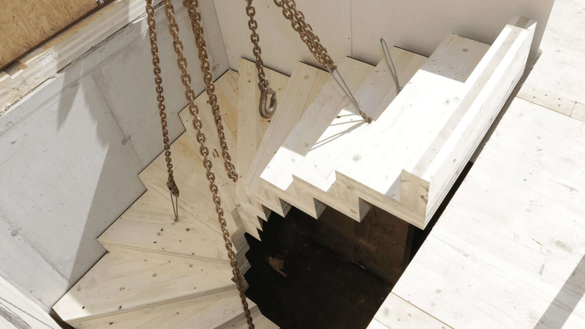 Holztreppe im Bau, über der Treppe hängen Ketten und Haken. Die Treppe ist teilweise in eine Beton- und Sperrholzkonstruktion eingebaut, Werkzeuge und Materialien sind vor Ort sichtbar.