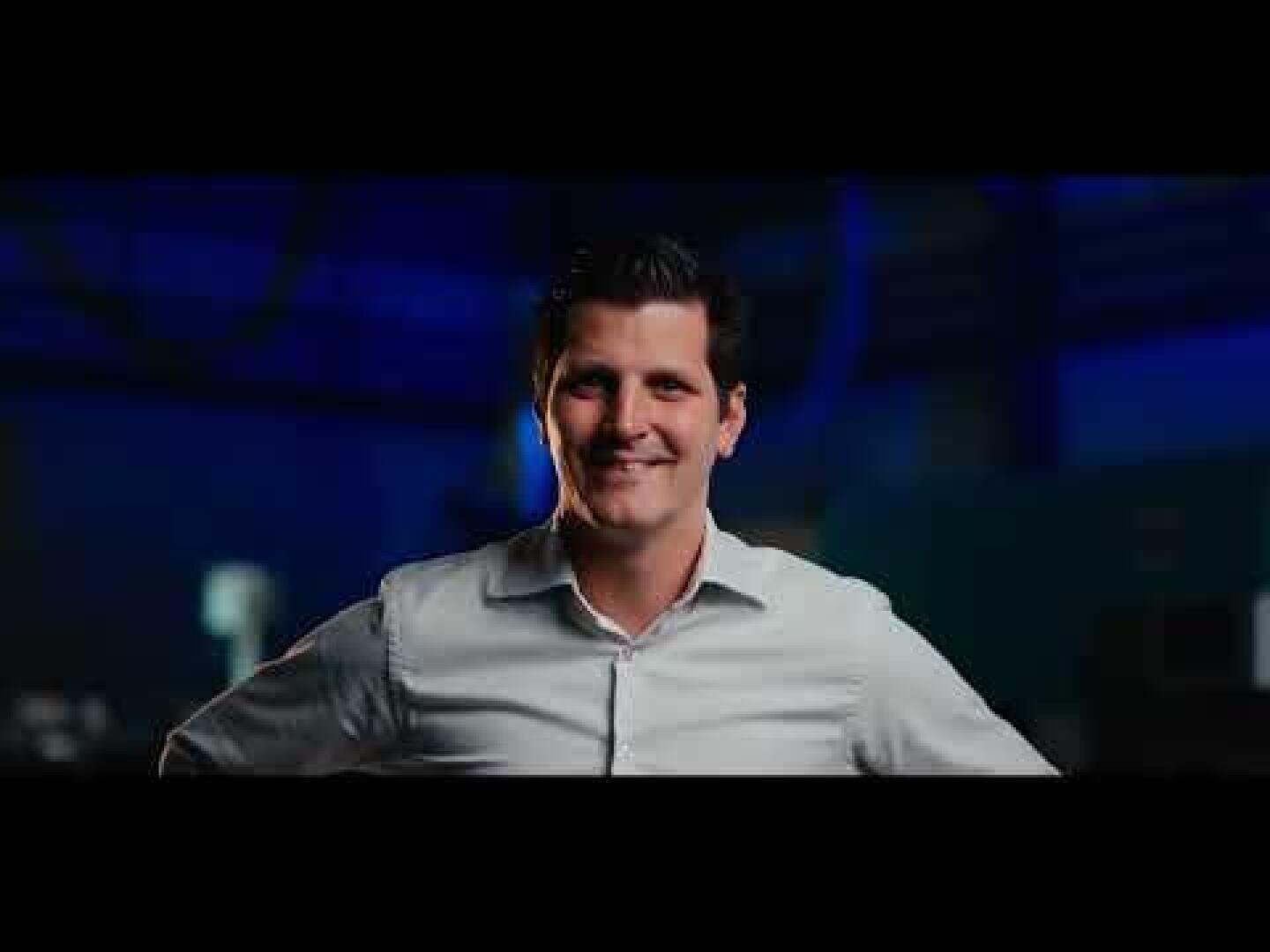 Ein Mann mit dunklem Haar, der ein helles Button-up-Hemd trägt, steht lächelnd mit den Händen in den Hüften vor einem verschwommenen, dunkelblauen Hintergrund.