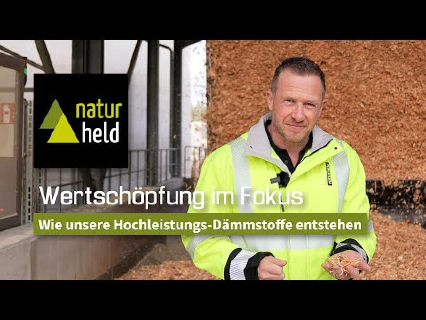 Ein Mann in einer neonfarbenen Sicherheitsweste hält Holzspäne vor einer Anlage. Der Text lautet: "naturheld Wertschöpfung im Fokus - Wie unsere Hochleistungs-Dämmstoffe entstehen.".