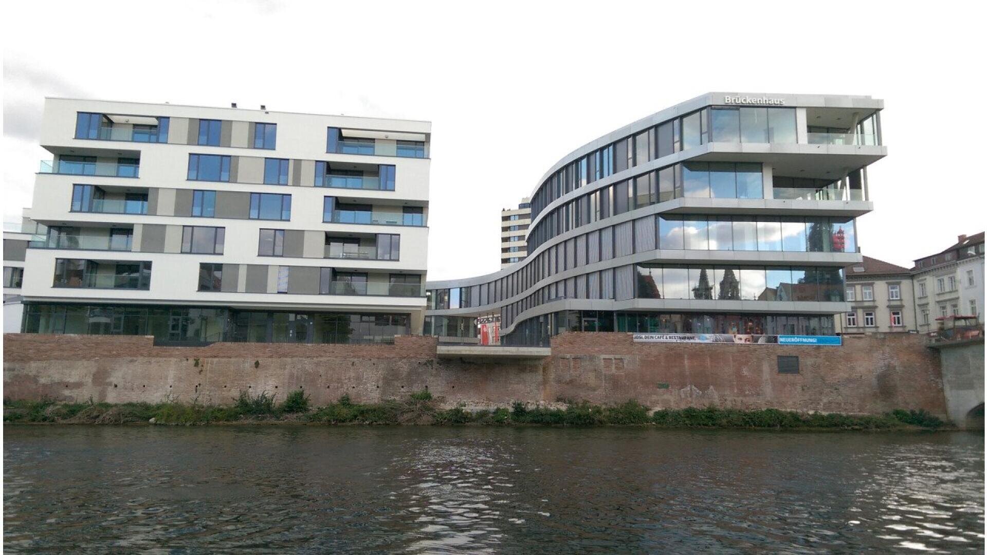 Modern, multi-story glass and white apartment buildings stand beside a river. The buildings have large windows and balconies, with a brick wall at the water’s edge. The sky is partly cloudy.
