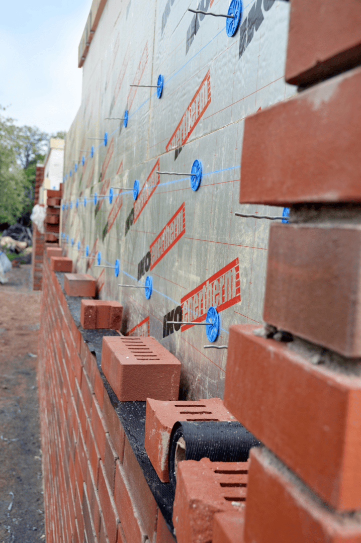 Nahaufnahme einer teilweise errichteten Ziegelwand mit Dämmplatten dahinter. Blaue Abstandshalter sind an den Paneelen befestigt, und lose Ziegel sind auf der Wand gestapelt. Im Hintergrund sind Baumaterialien zu sehen.